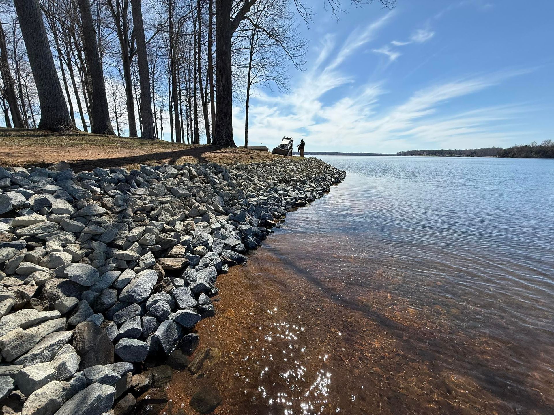 Rocky lakeshore with bare trees, clear water, and two people in the distance under a blue sky
