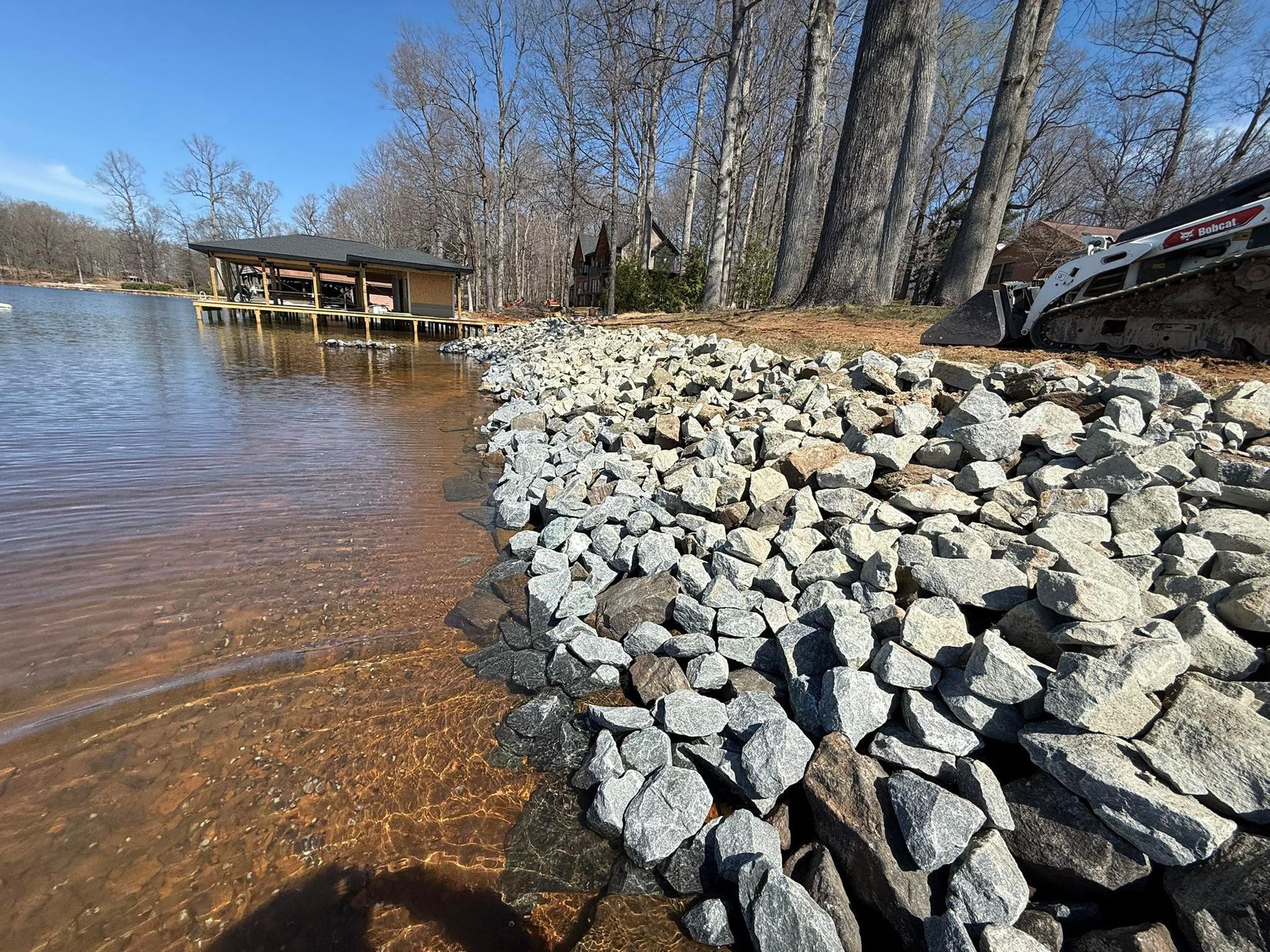 Rock-lined shoreline beside a calm lake with a wooden dock and leafless trees under a clear blue sky
