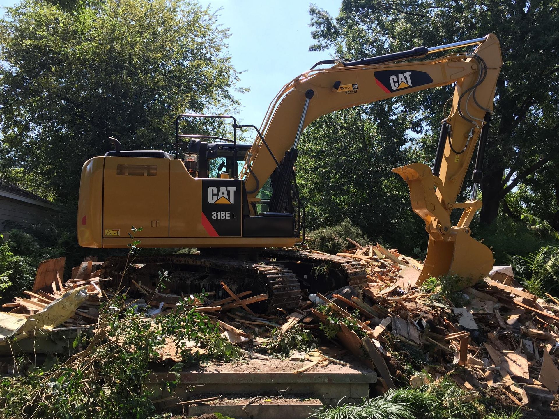 Yellow CAT excavator demolishing a house, debris pile, trees in the background, sunny day.