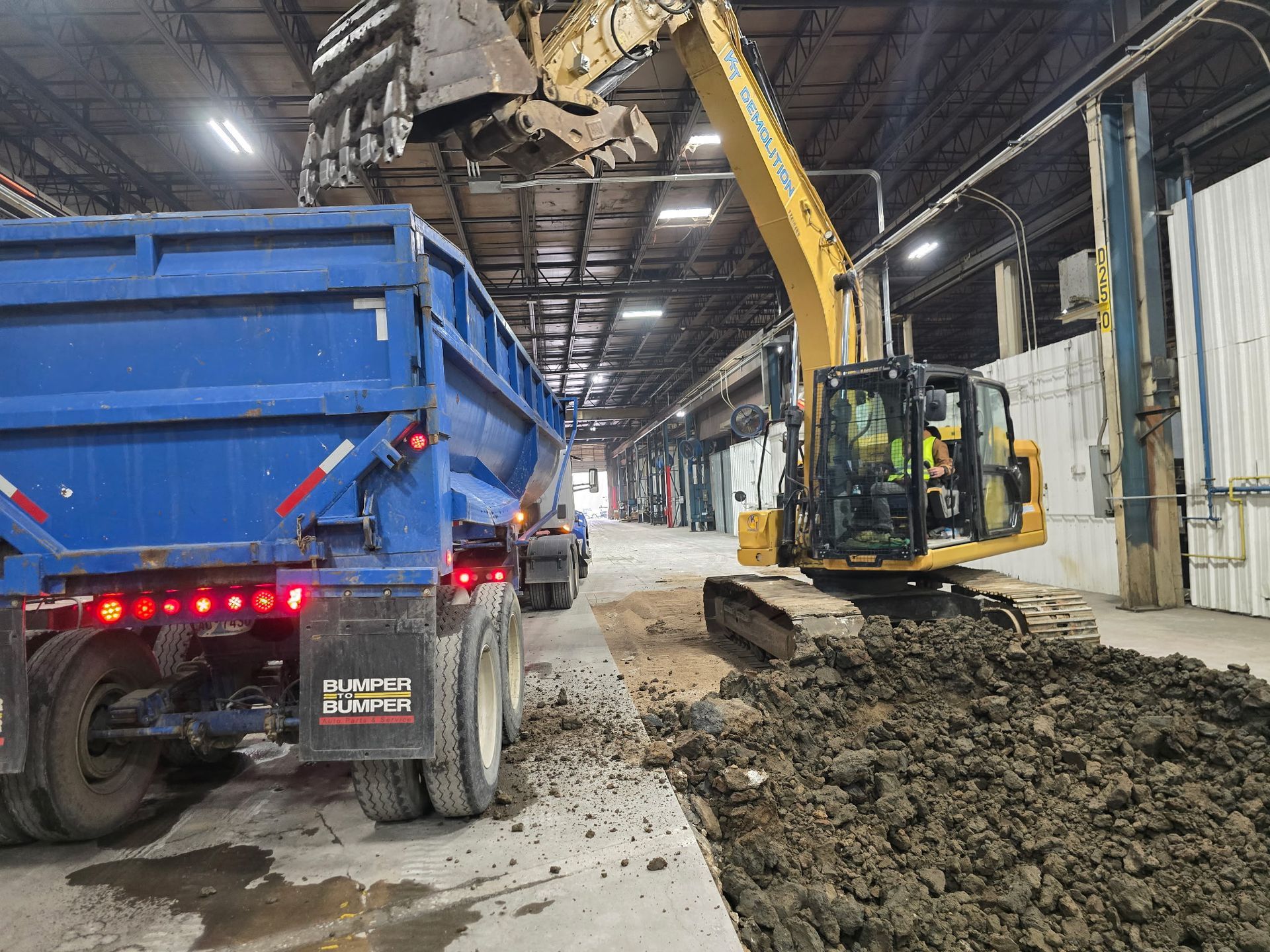 Excavator loading dirt into a blue dump truck inside a building.