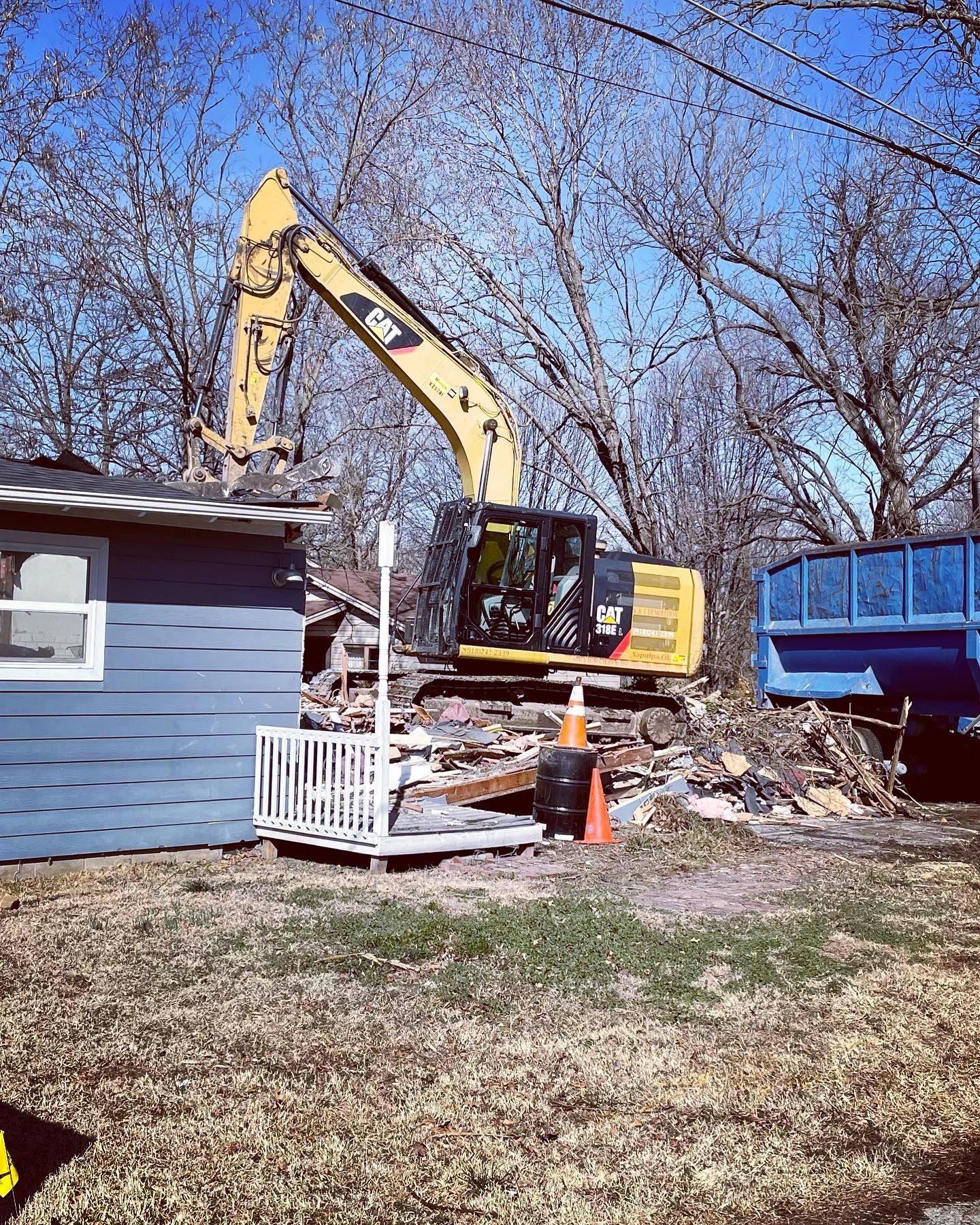 Yellow excavator demolishing a small blue building on a sunny day. Debris and a blue dumpster nearby.