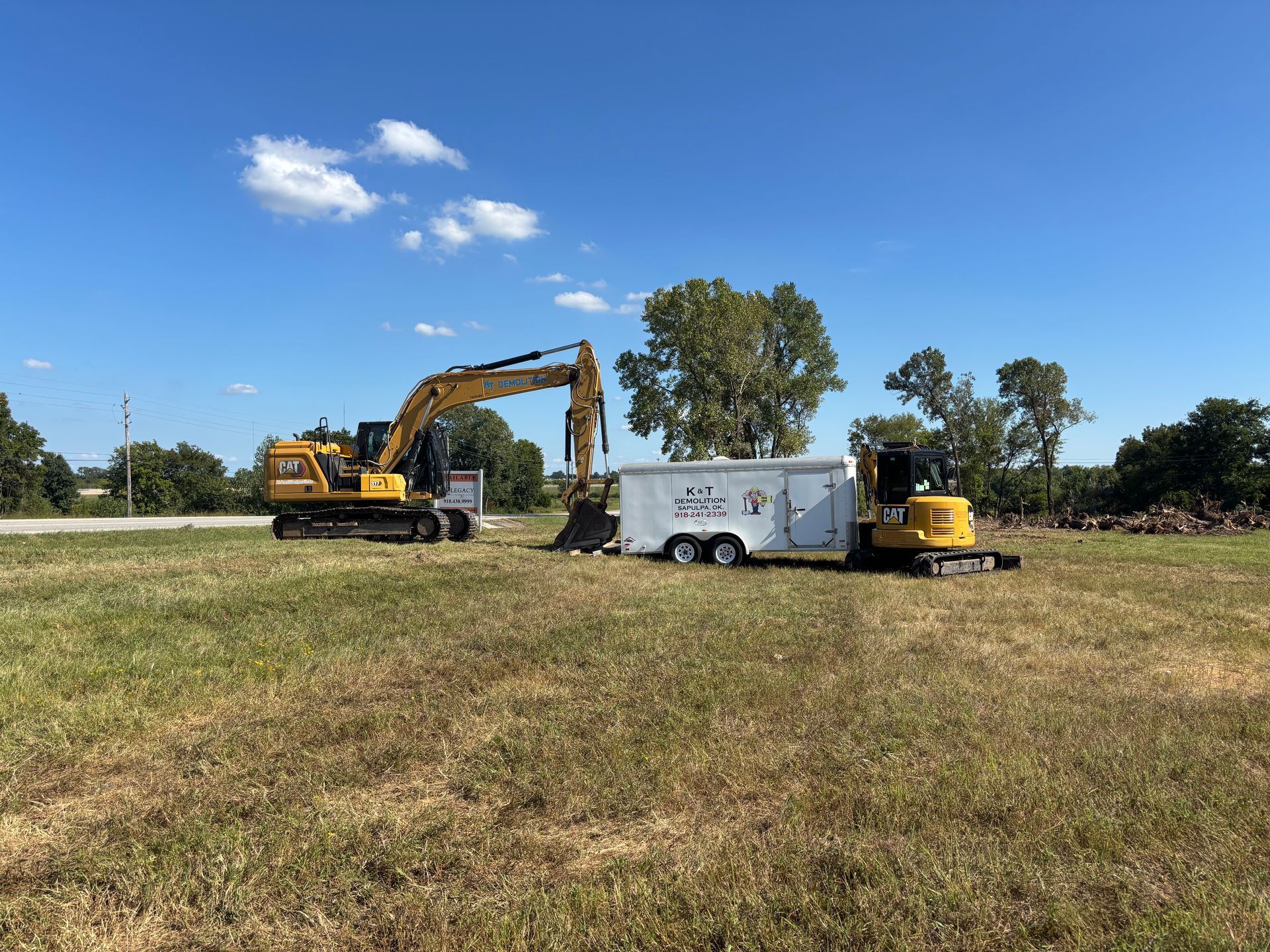 An excavator and mini-excavator work near a white trailer in a grassy field under a blue sky.
