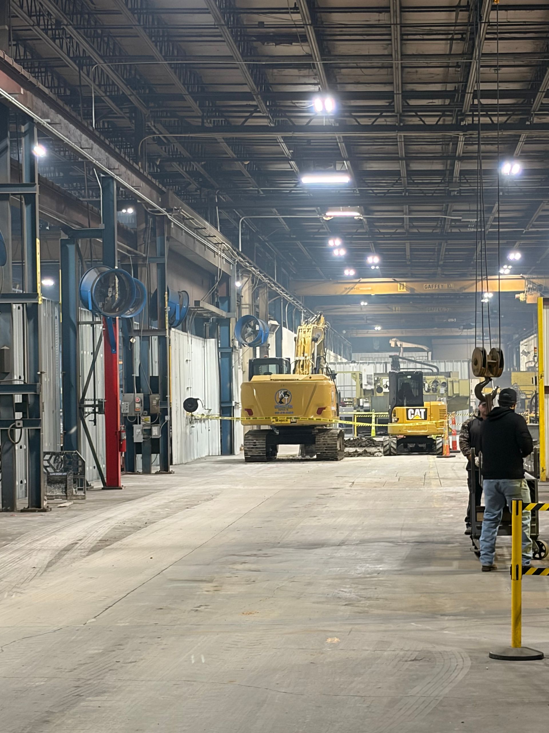 Large factory interior with heavy machinery, including an excavator. Two workers walk in the foreground.