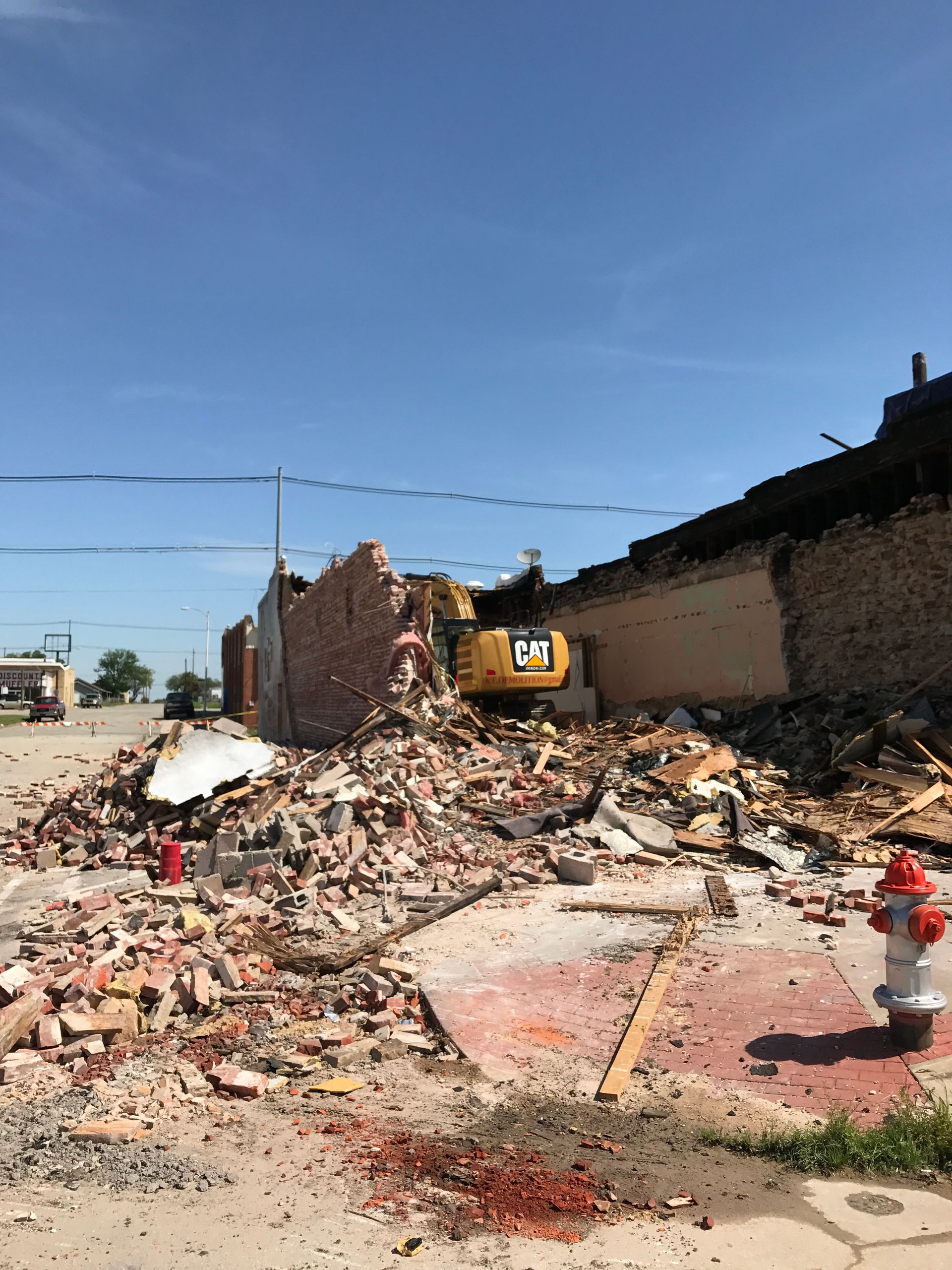 Demolition of a brick building by a yellow excavator on a sunny day. Rubble covers the ground.