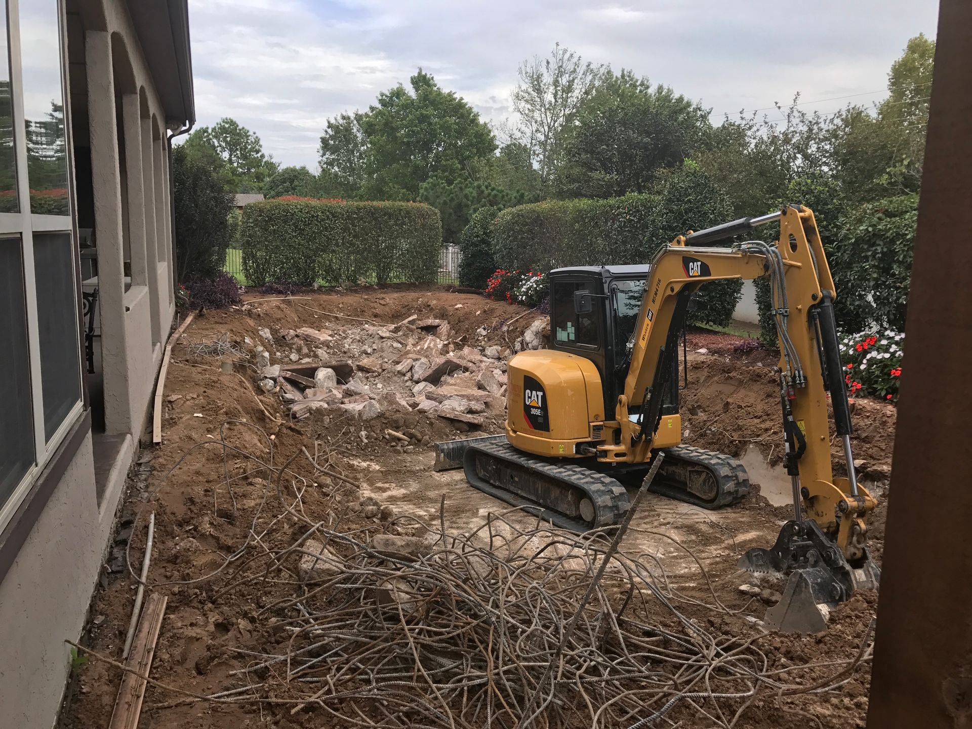 Yellow excavator demolishing a yard, near a building and hedges.