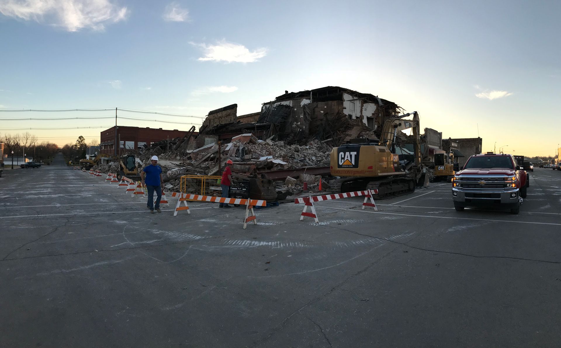 Demolished building with heavy machinery and debris, street barricades, clear sky.