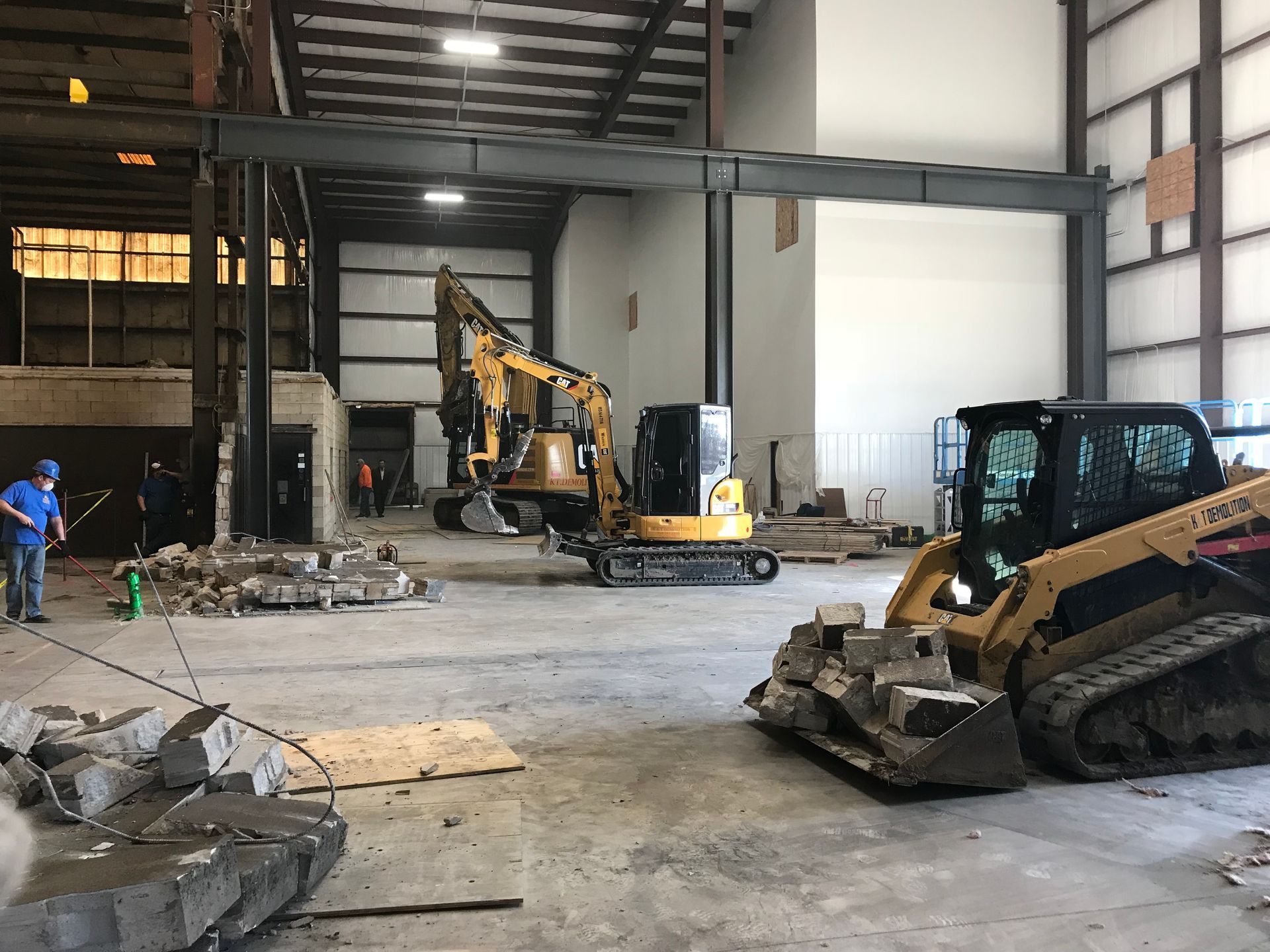 Construction site with heavy machinery inside a warehouse. One worker, debris, and structural steel beams visible.