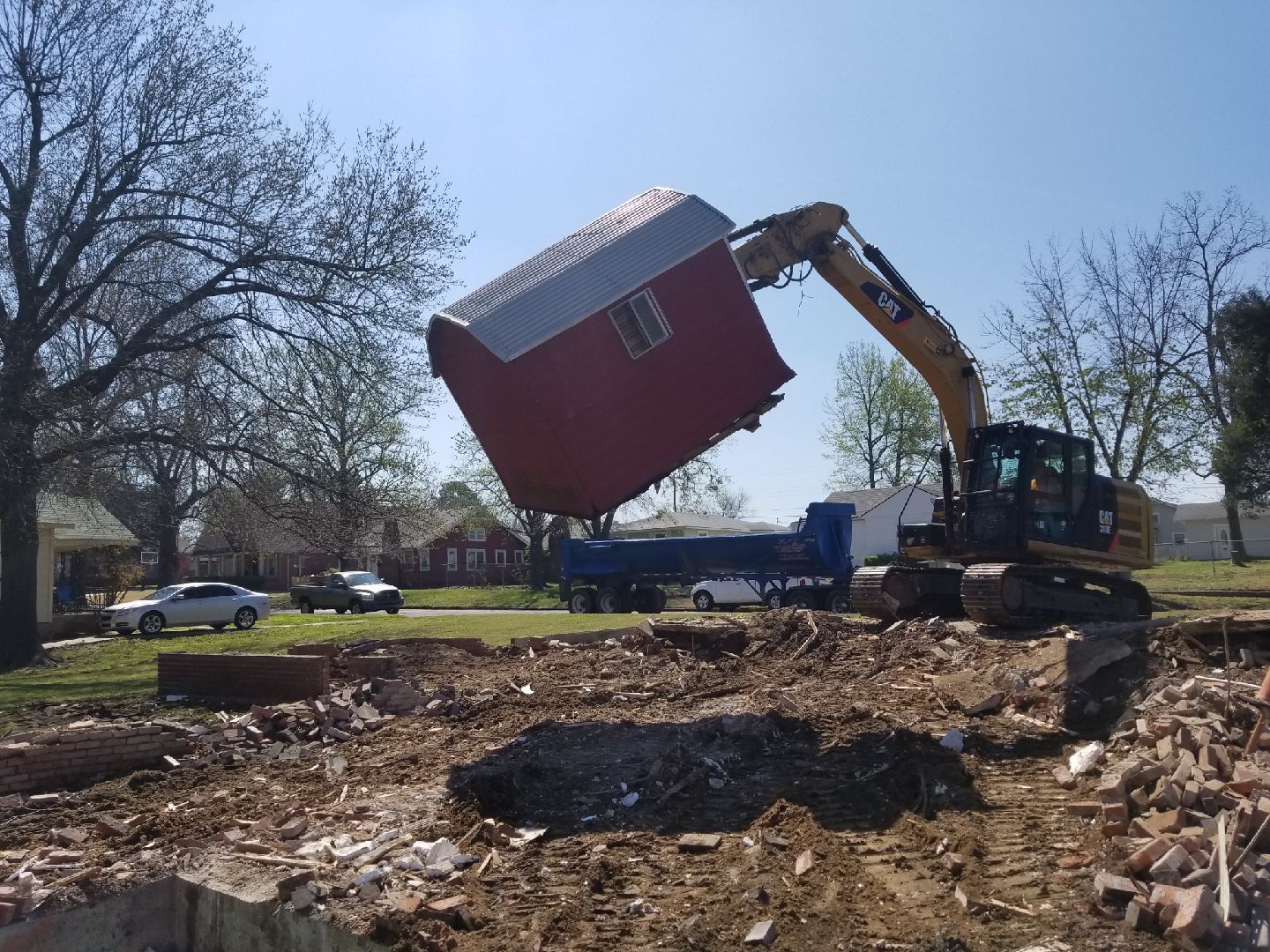 Excavator lifting a red barn structure over a construction site.