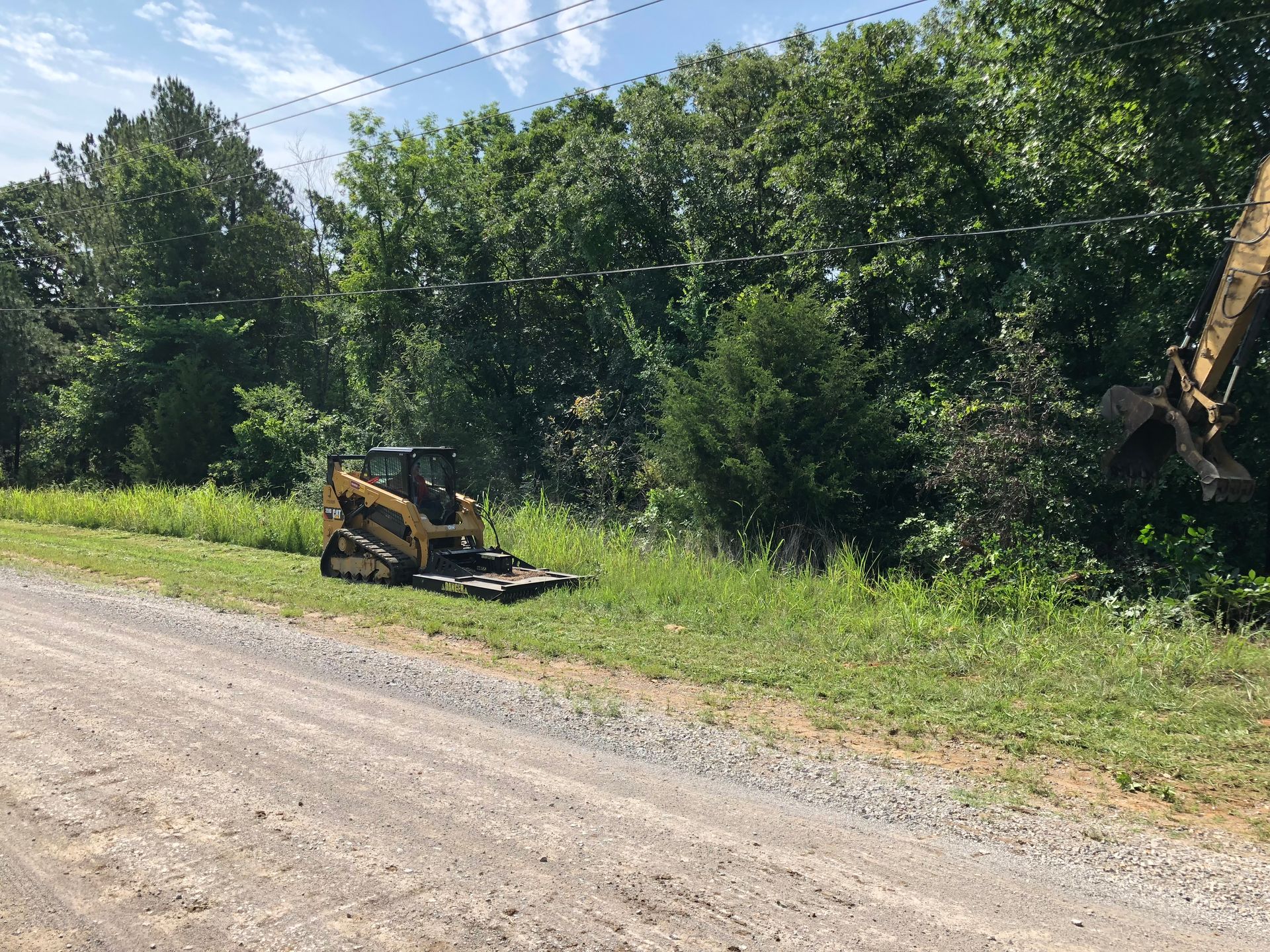 A skid steer and excavator clearing brush alongside a gravel road near trees under a bright sky.