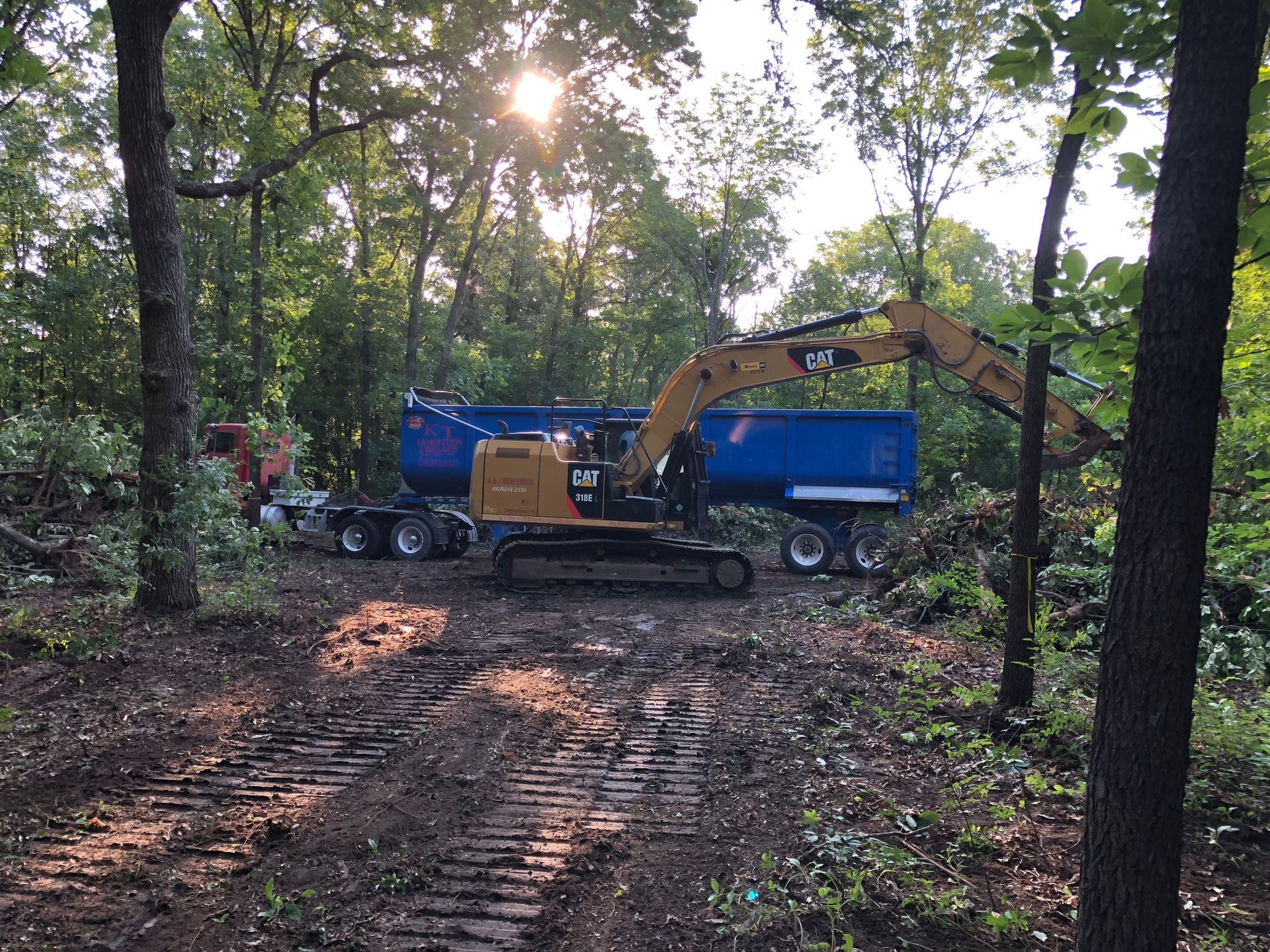 Excavator loading a blue dump trailer in a wooded area. Sunlight shines through the trees.