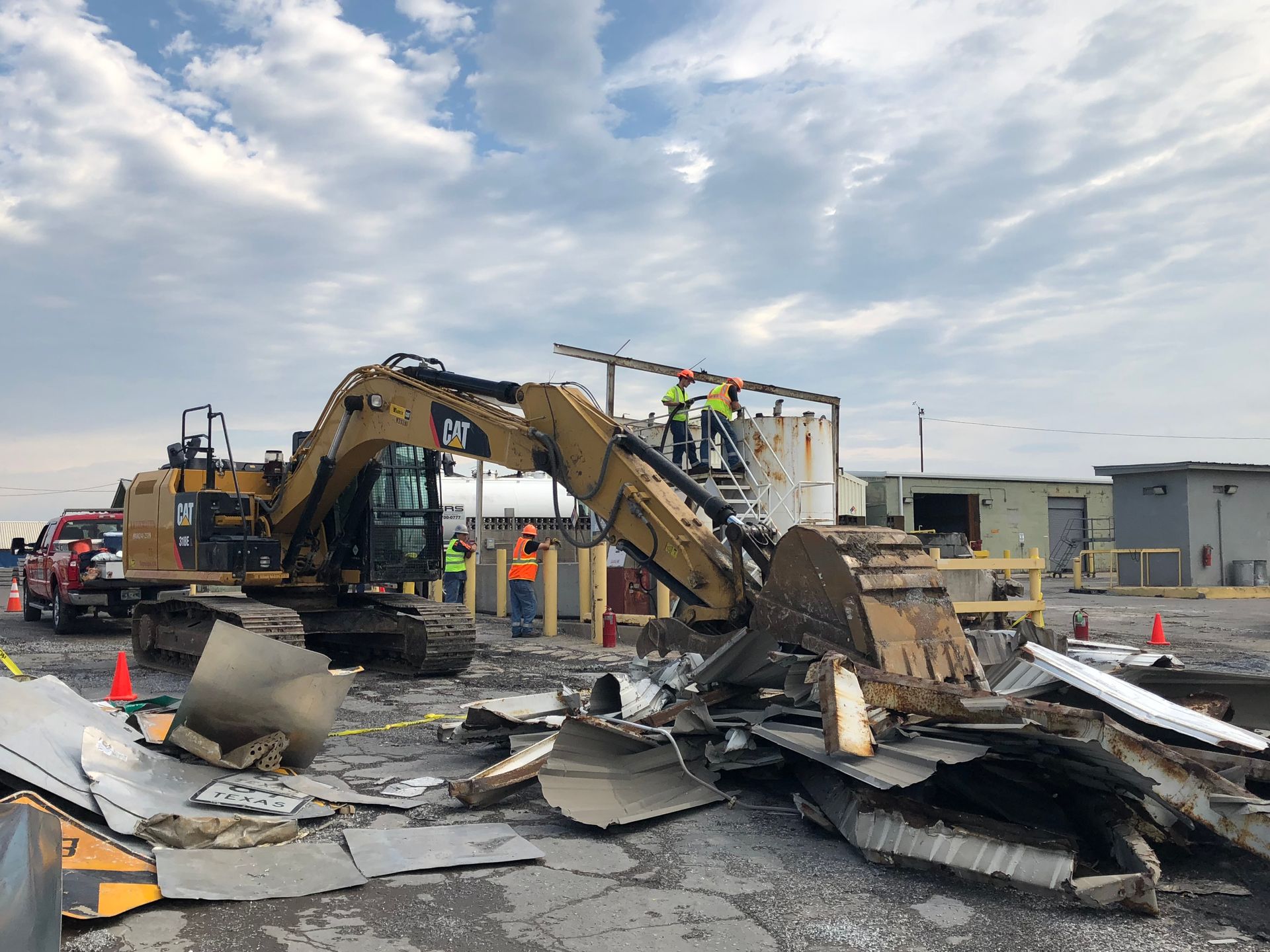 An excavator demolishes a structure in a construction zone; workers in safety vests observe, cloudy sky overhead.