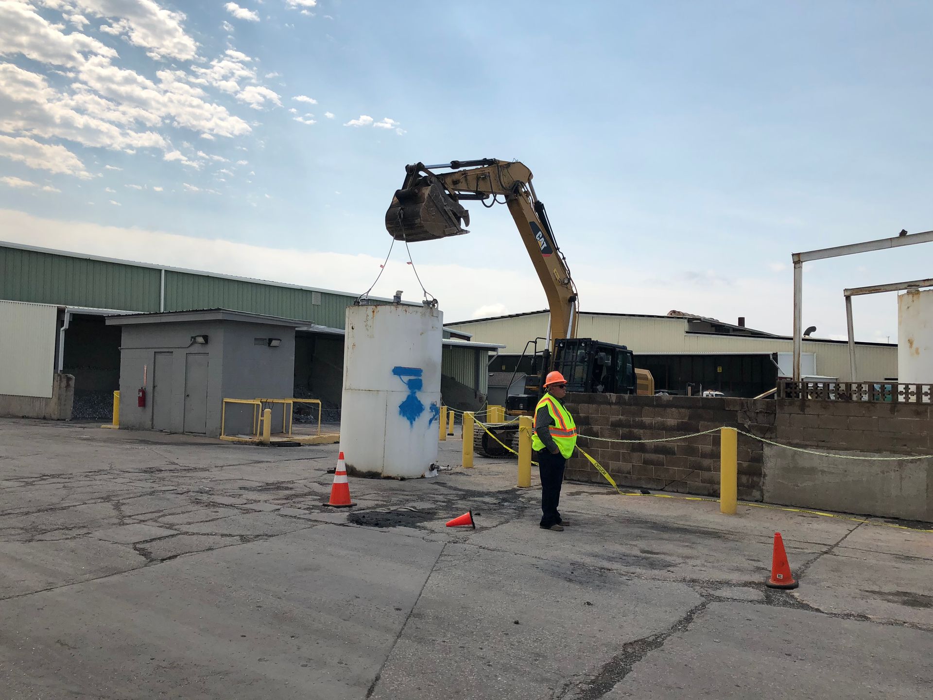 An excavator lifting a large white container with a worker in a safety vest watching in an industrial setting.