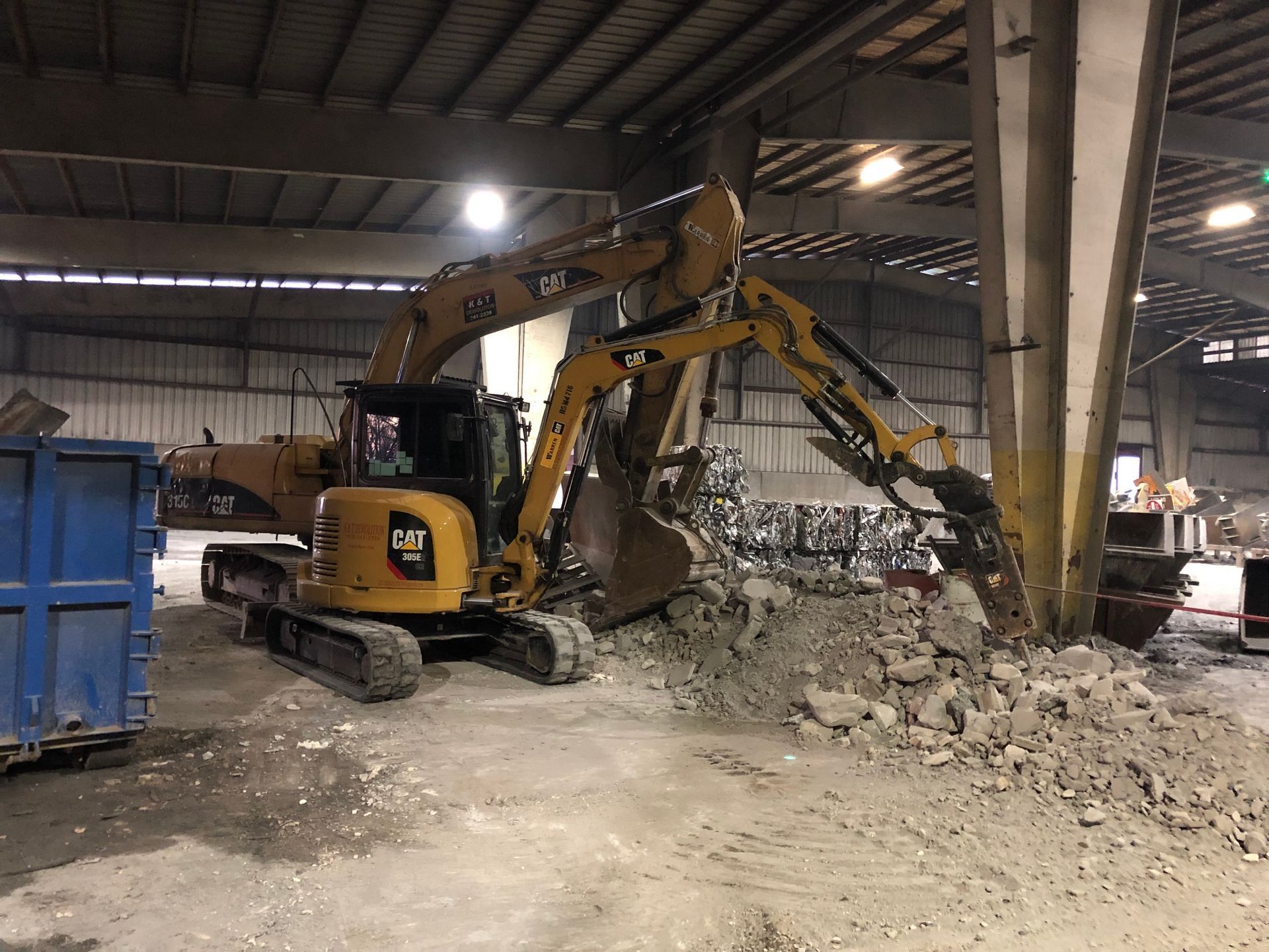 A yellow Caterpillar excavator scoops up debris inside a large warehouse.