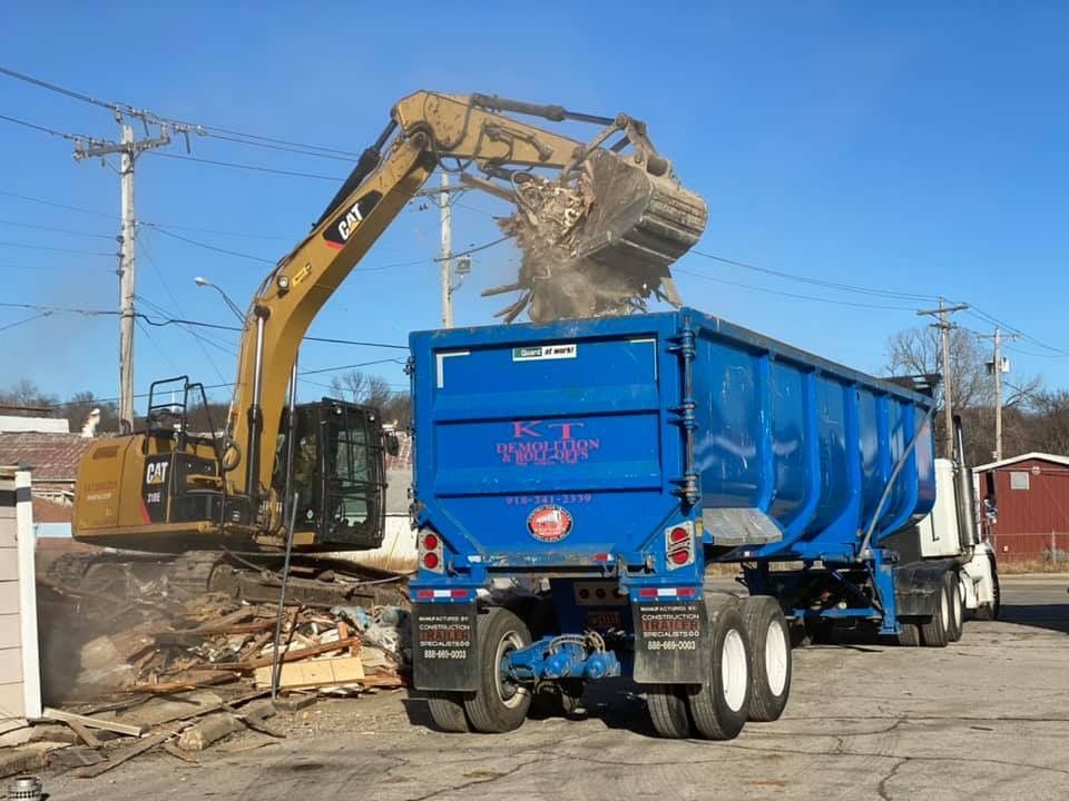 Excavator loading debris into a blue dump trailer in an outdoor setting.