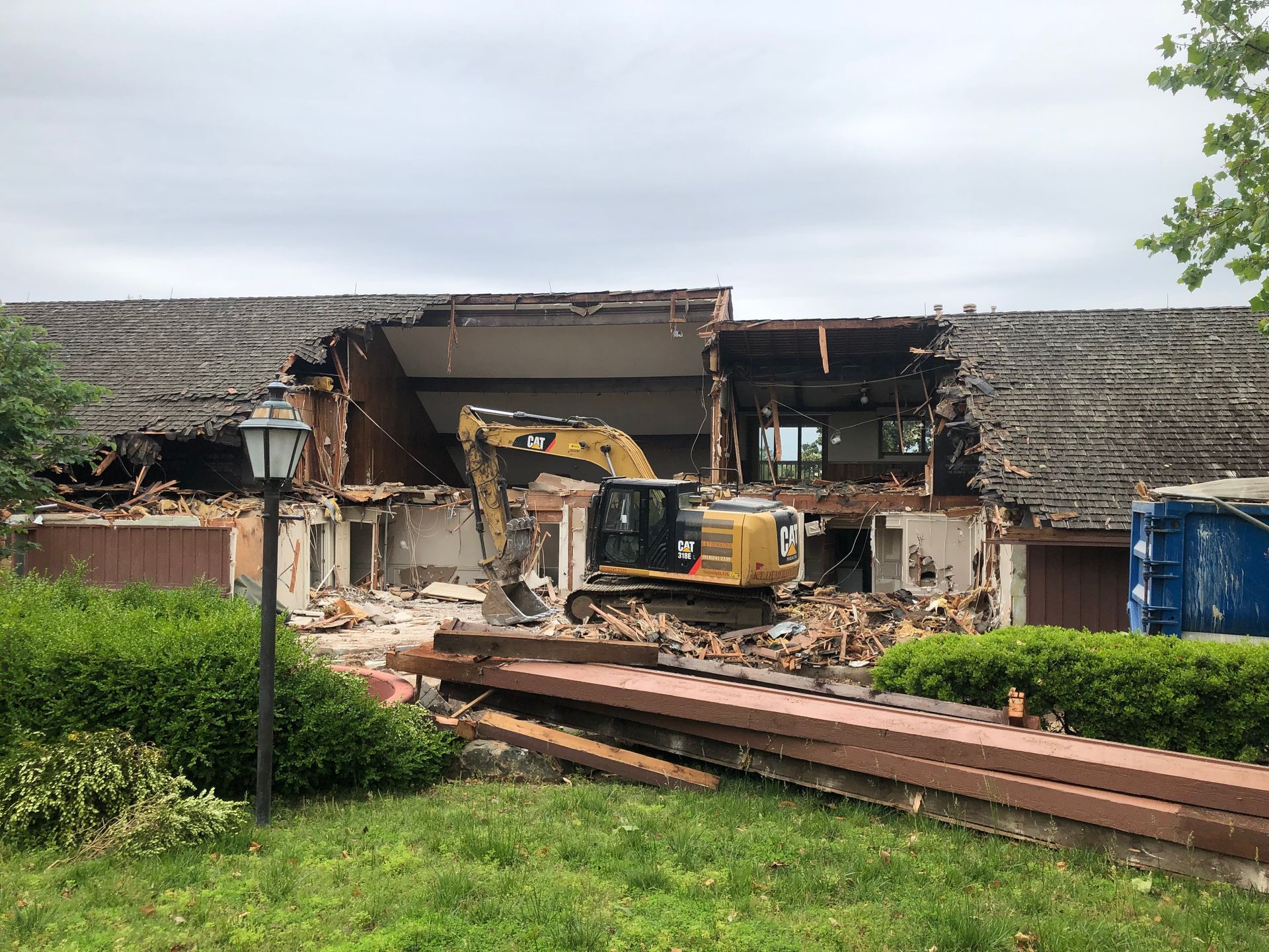 Demolition of a building. Yellow excavator tears into a structure with brown roof. Green grass and blue container in view.