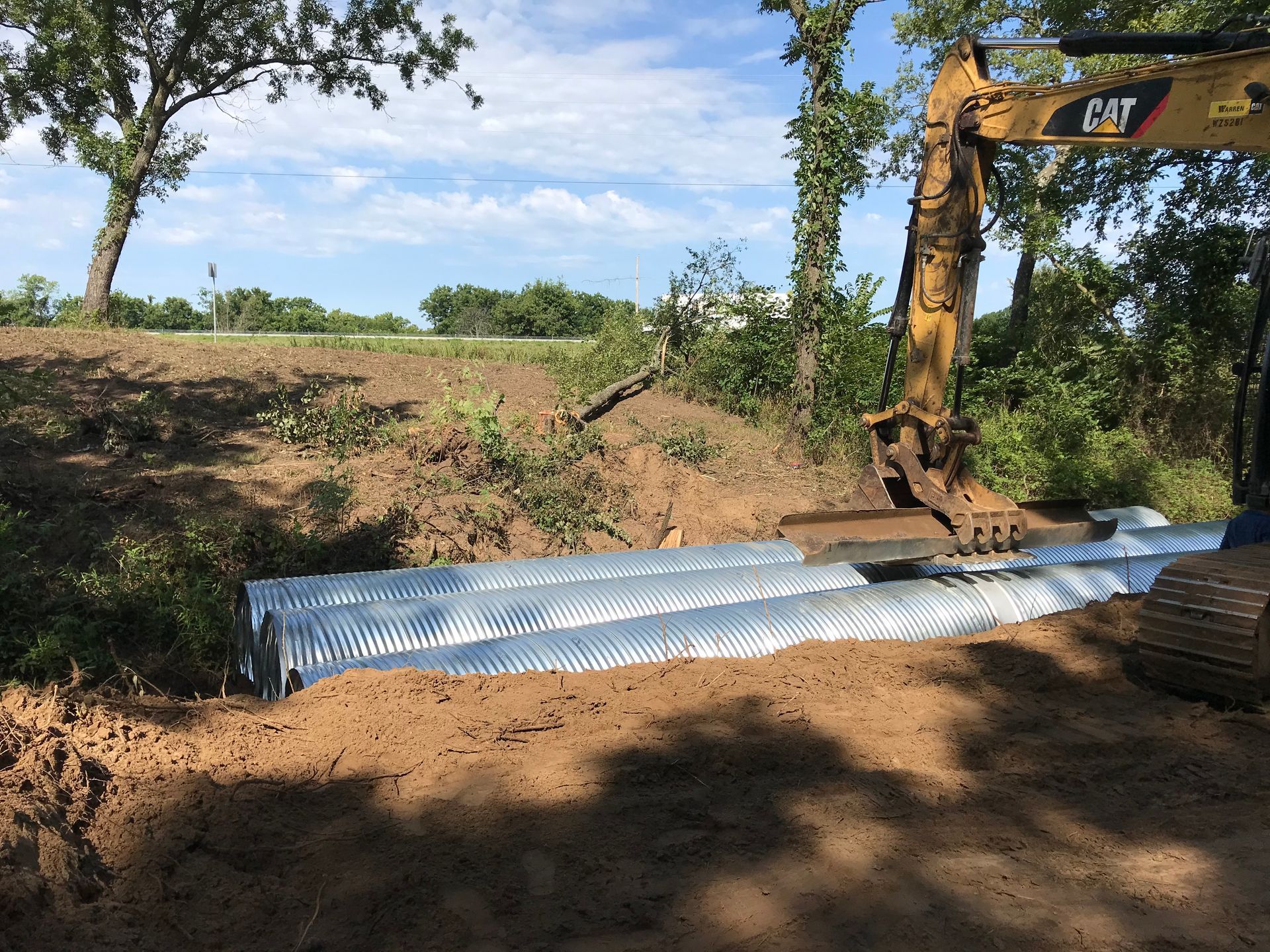Excavator placing corrugated metal pipes in a ditch for road construction.