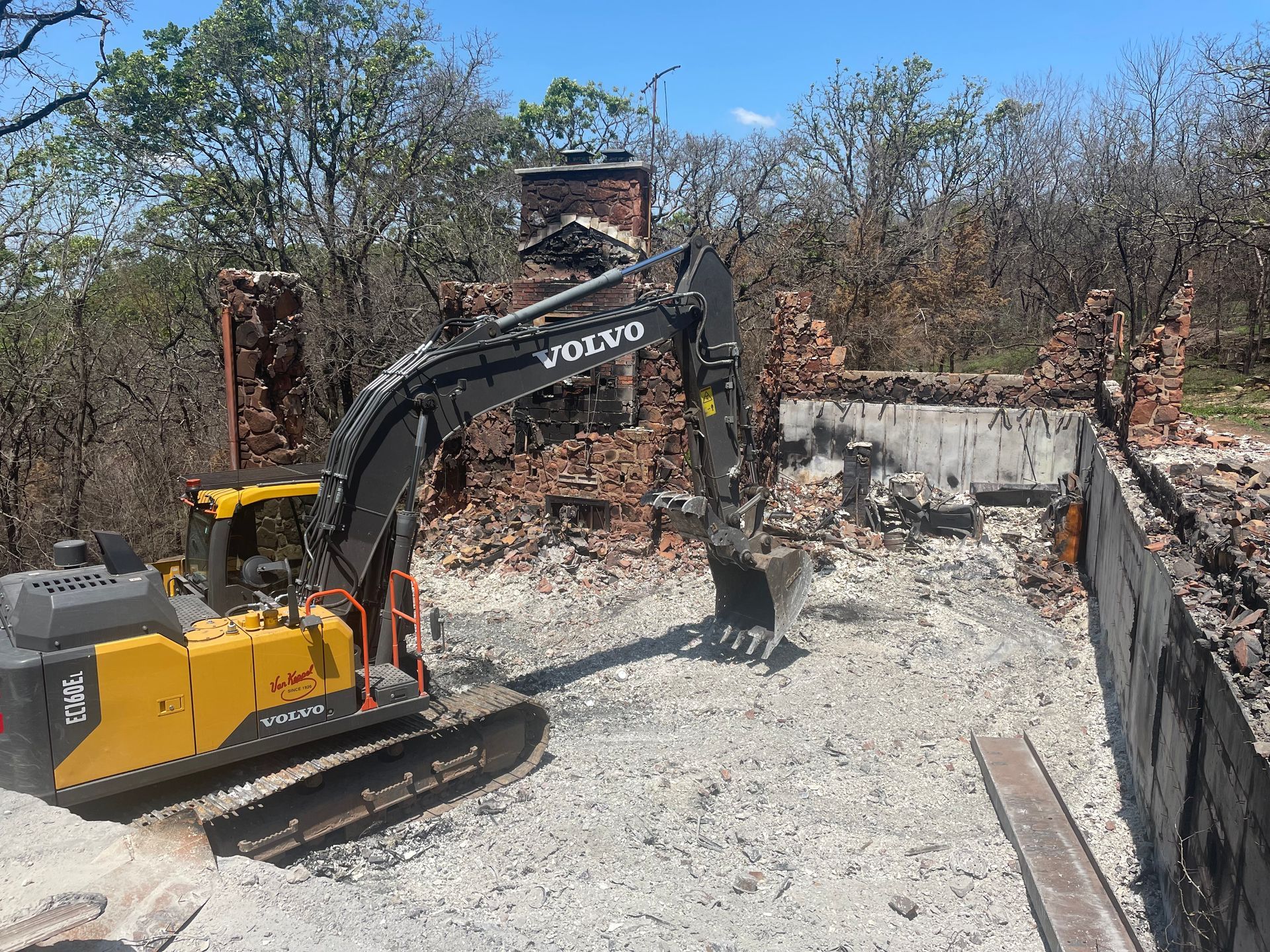 Yellow Volvo excavator demolishing a brick structure, trees in the background.