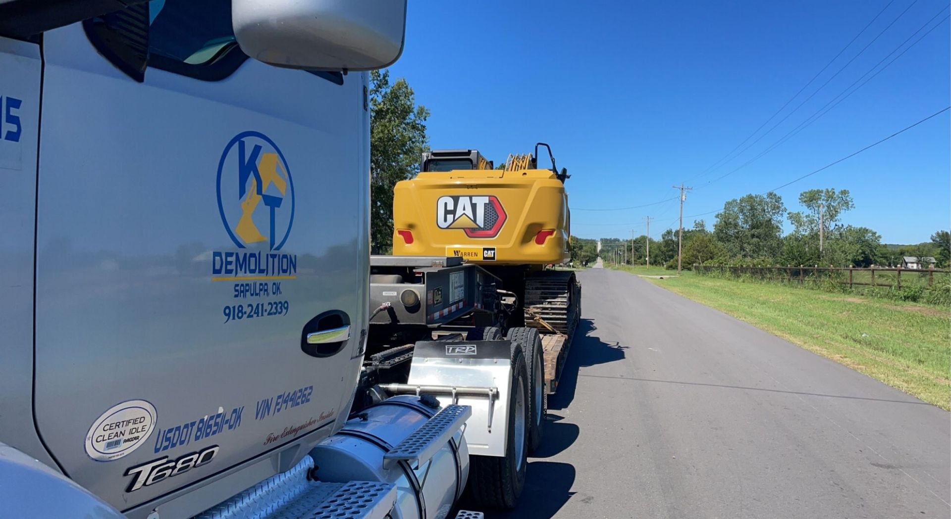 A semi-truck hauling a yellow CAT excavator down a road on a sunny day.