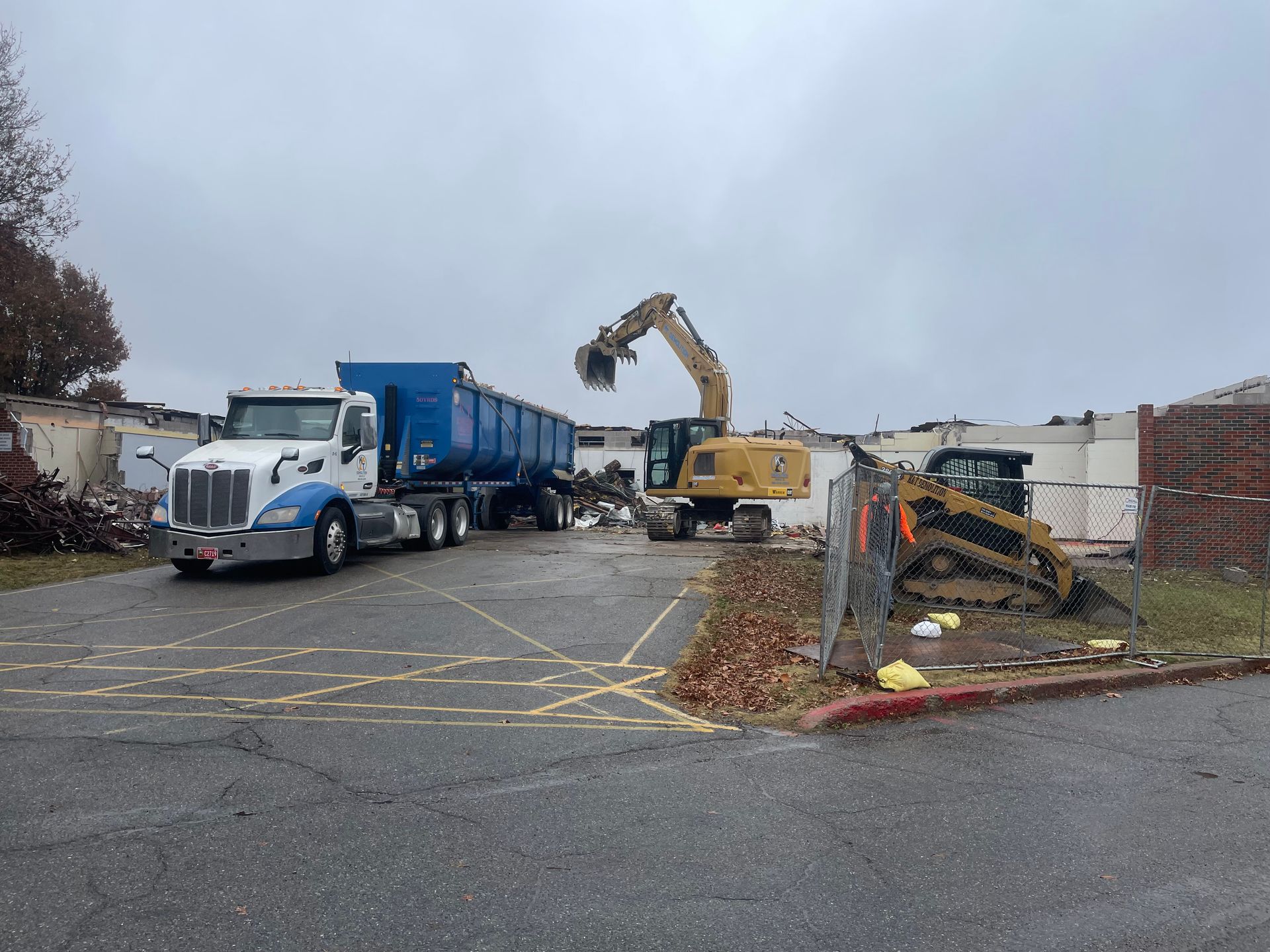 Demolition site with truck, excavator, and skid steer clearing debris on a cloudy day.