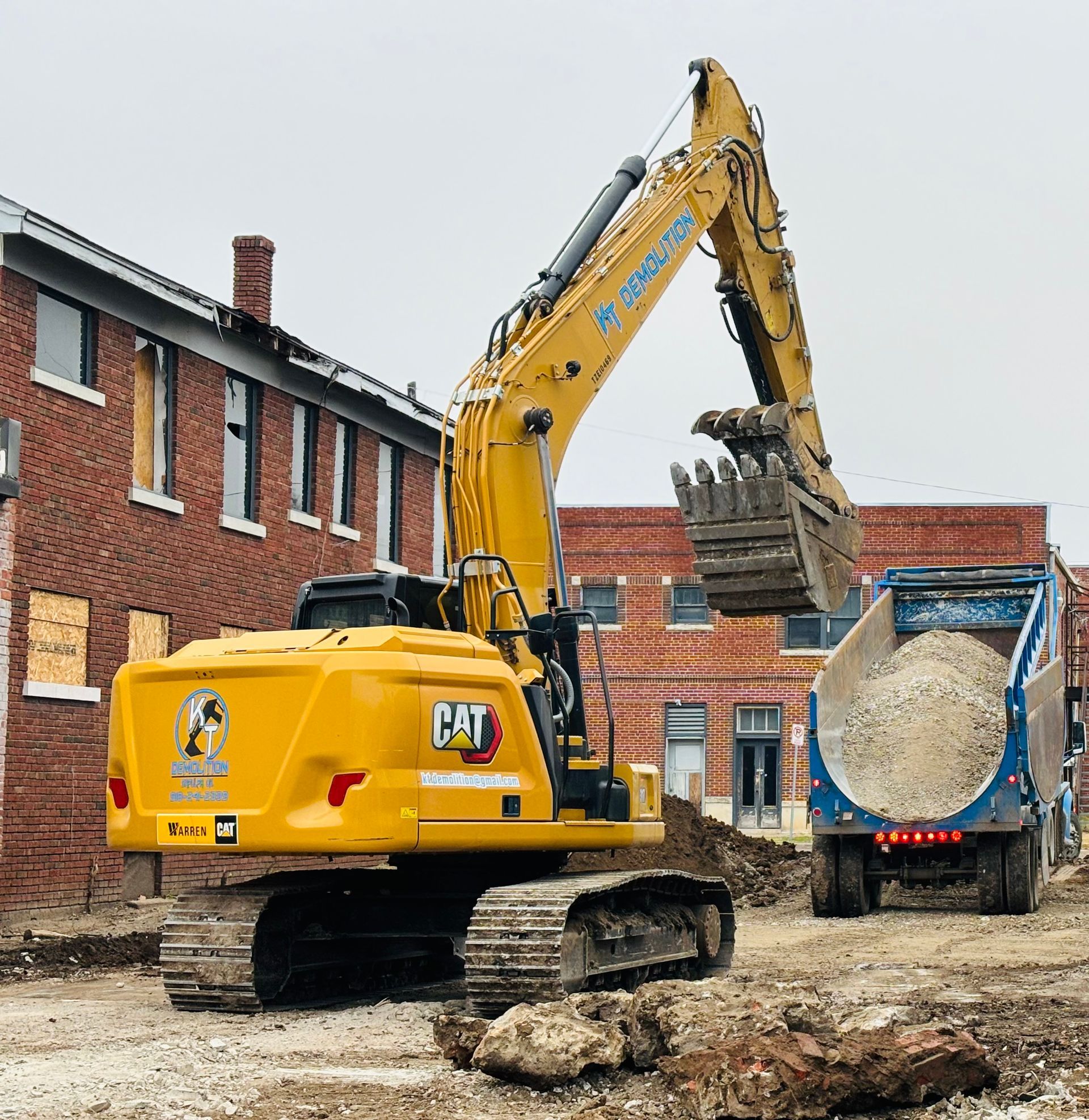 Yellow excavator loading gravel into a blue dump truck next to a brick building.