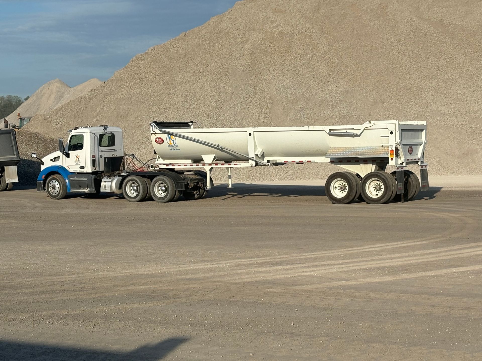 White semi-truck with a long trailer parked near a large pile of gravel in a quarry setting.