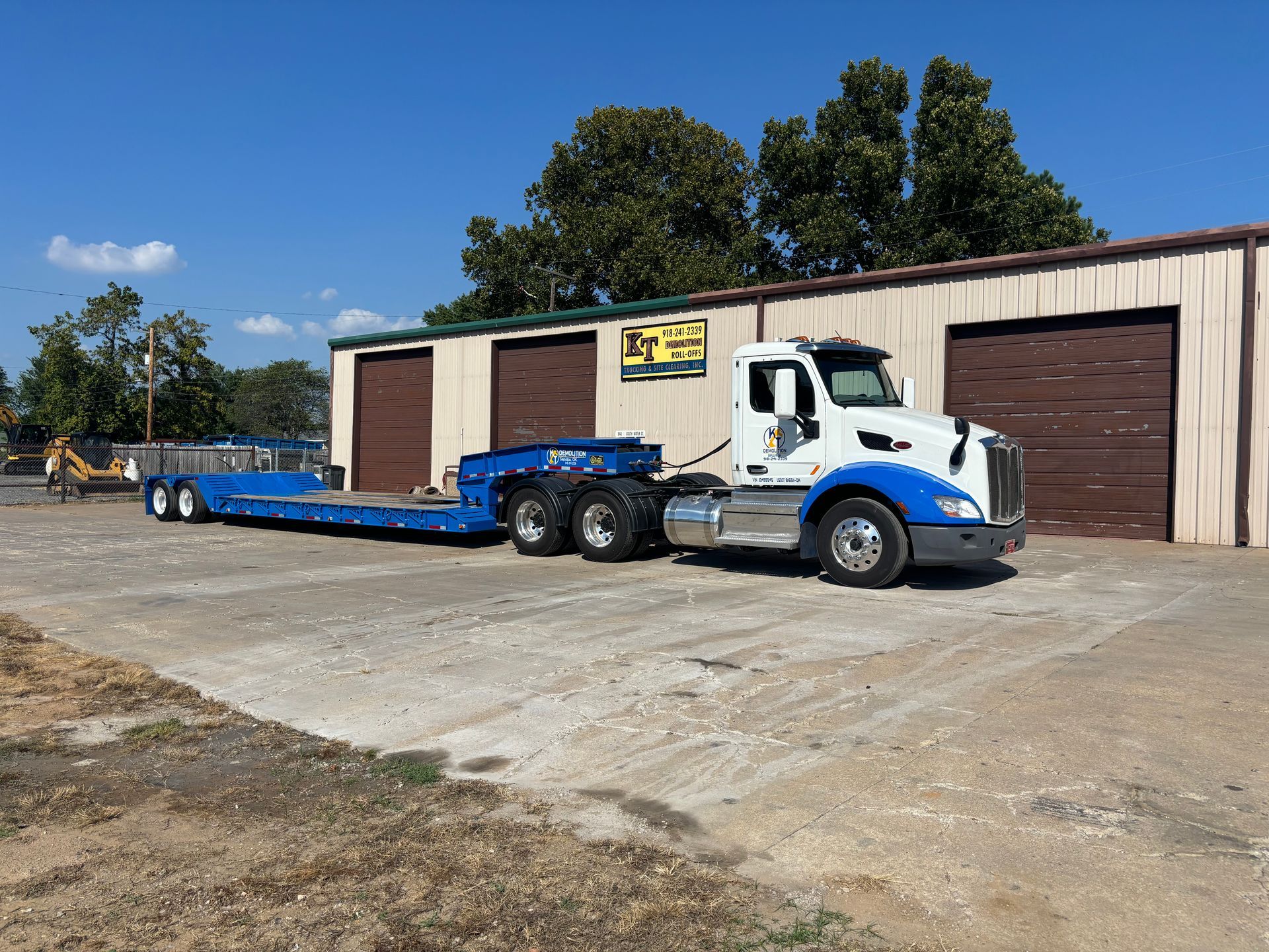 White and blue semi-truck with a flatbed trailer parked in front of a building on a sunny day.