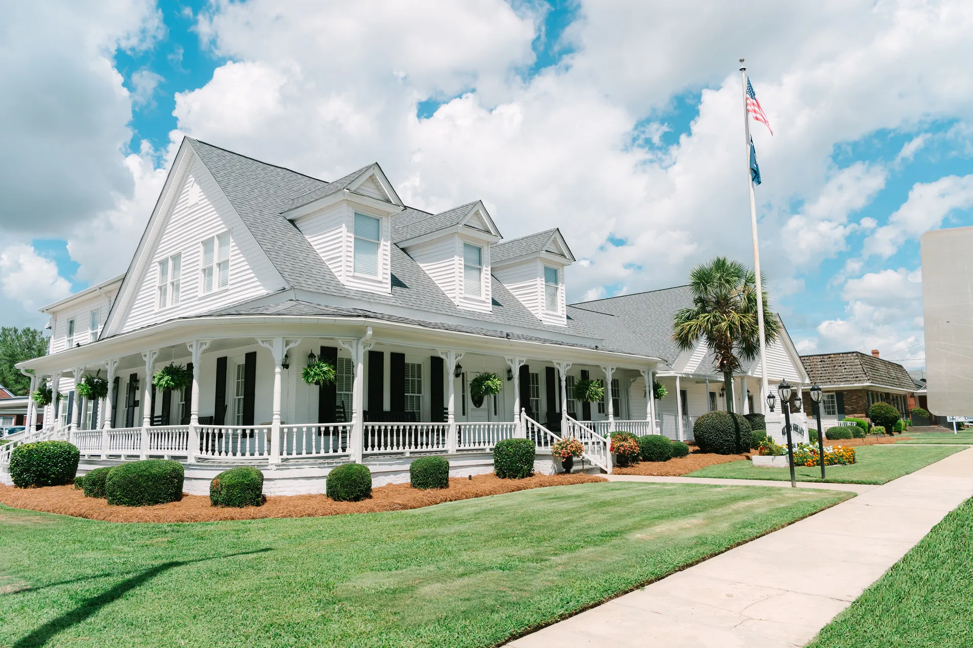 A large, two-story white house with a wrap-around porch, multiple gabled roof dormers, and a manicured lawn under blue sky.