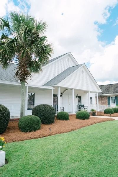 A white suburban house with a gray roof, a palm tree in the front yard, and neatly trimmed shrubs along the porch.