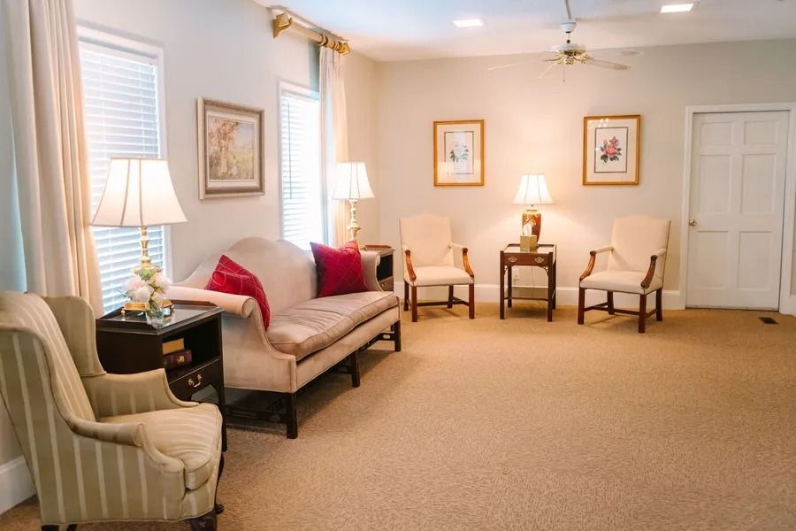 A bright, neutral-toned living room featuring a sofa with red pillows, three armchairs, lamps, and wall art.