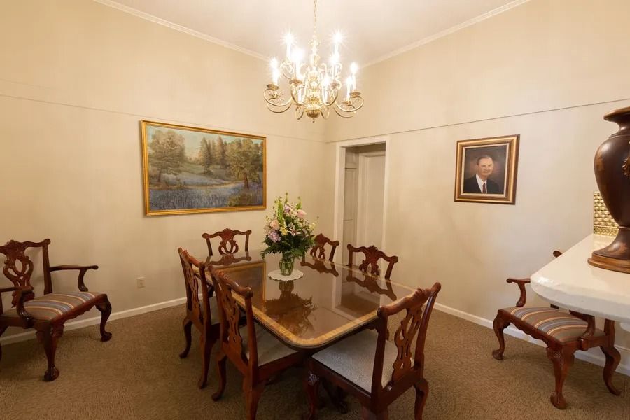 A dining room with a glass-top table, six ornate chairs, a chandelier, wall art, and a small portrait on a beige wall.