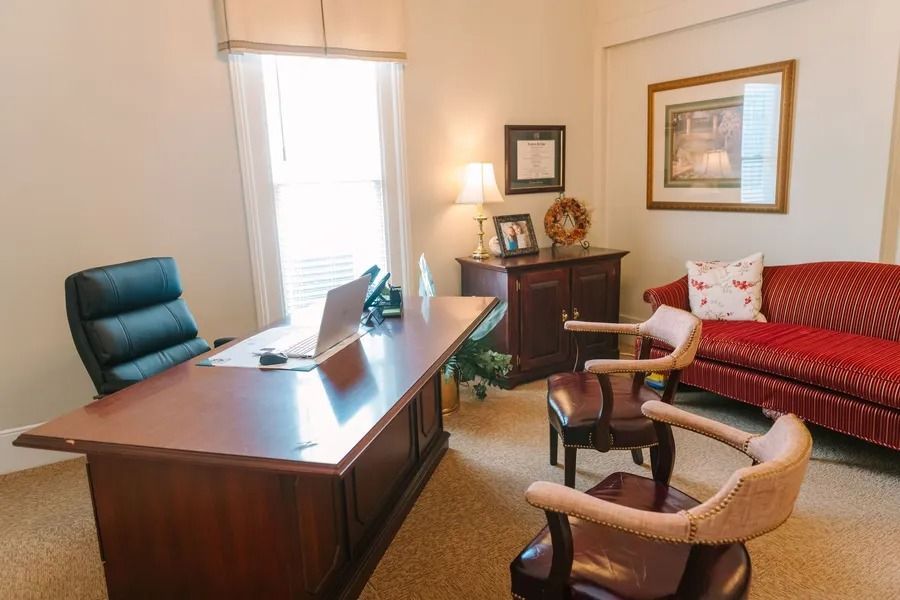 A professional office with a wooden desk, dark chair, two armchairs, a red sofa, and decor on walls and a cabinet.