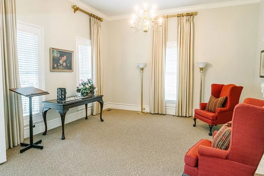 A formal room with beige carpet, two orange armchairs, a side table, a black lectern, and long curtains over windows.