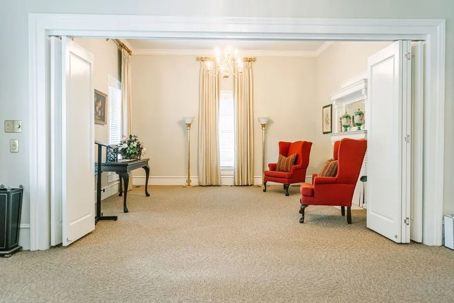 A brightly lit parlor with two red armchairs, beige carpet, white folding doors, and a small side table near a window.