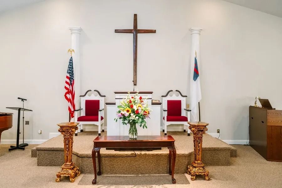A church altar with a wooden cross, two red chairs, an American flag, a white flag, and a floral centerpiece on a table.