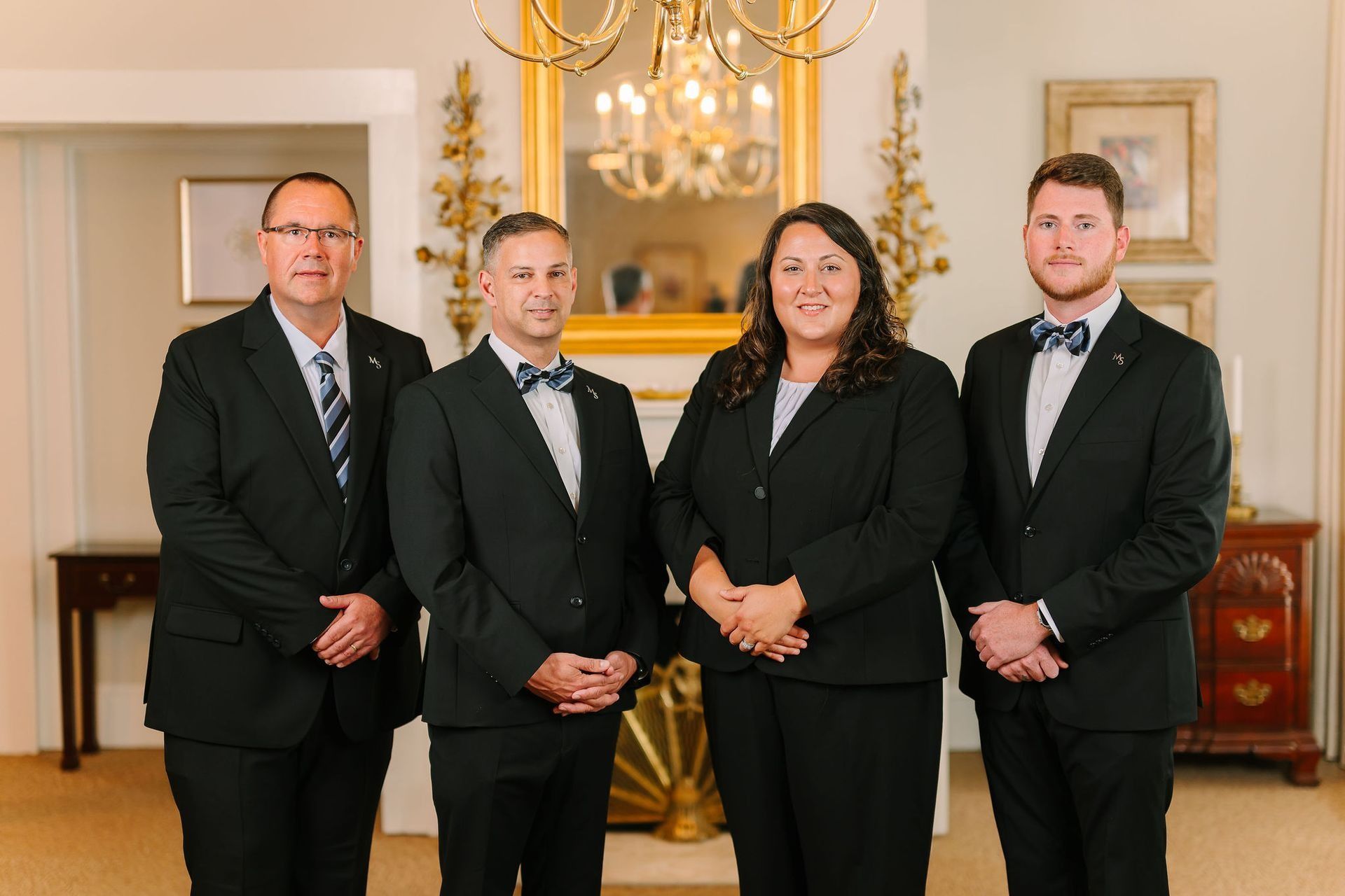 Four professionals in black suits stand in a line against an indoor backdrop with a fireplace and mirror.