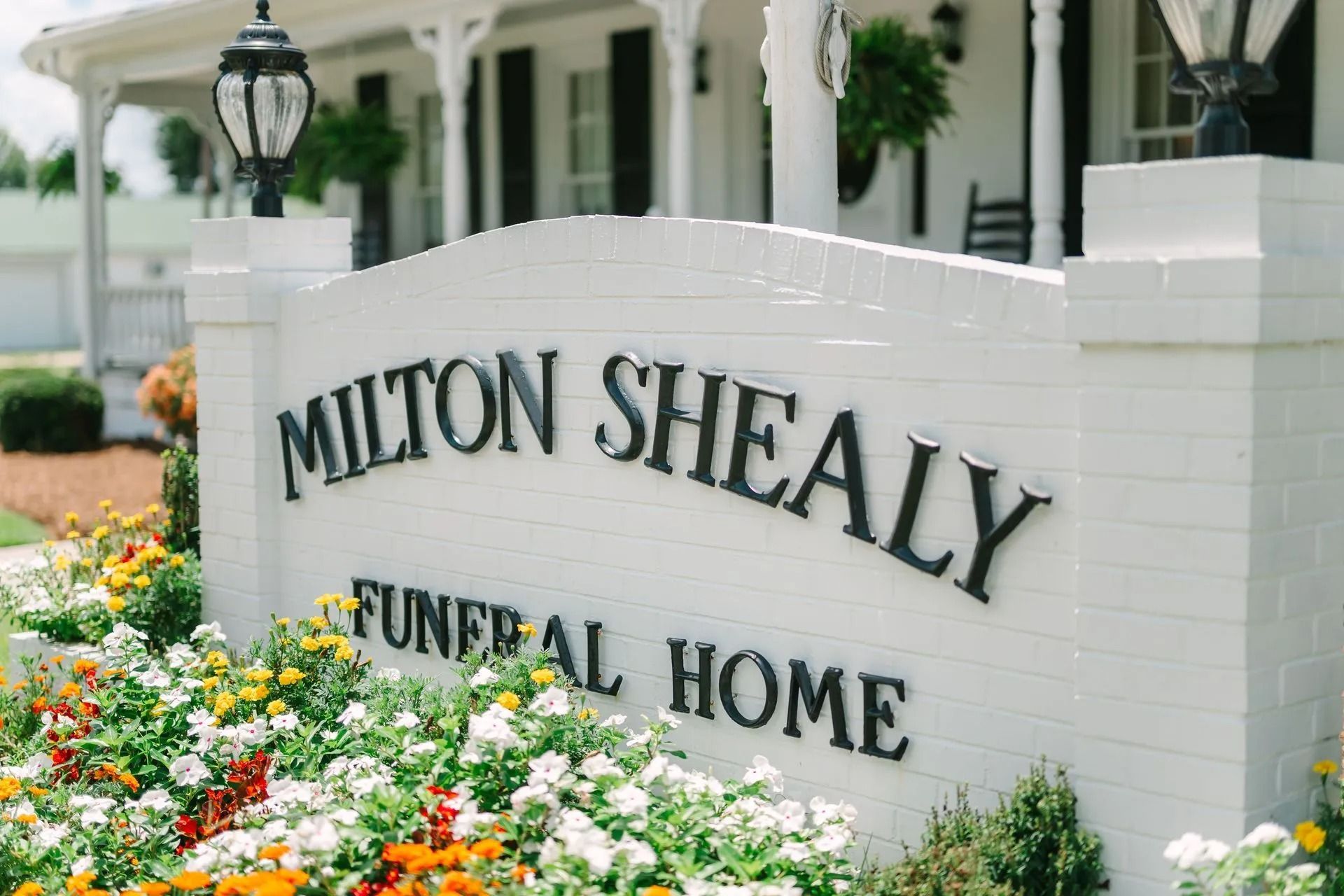 A white brick sign for Milton Shealy Funeral Home surrounded by colorful flowers, with a porch in the background.
