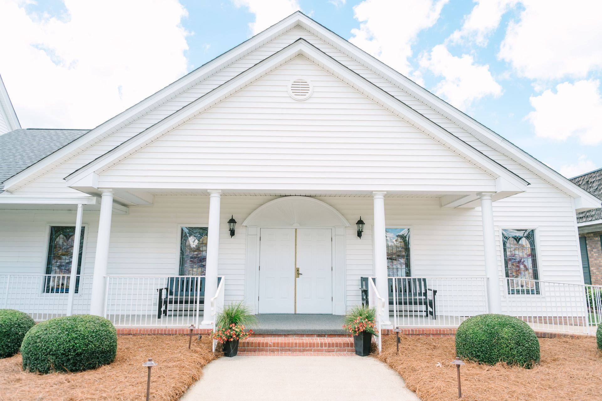 A white church with a peaked roof, arched double doors, stained-glass windows, and green shrubs on a wood-mulched lawn.