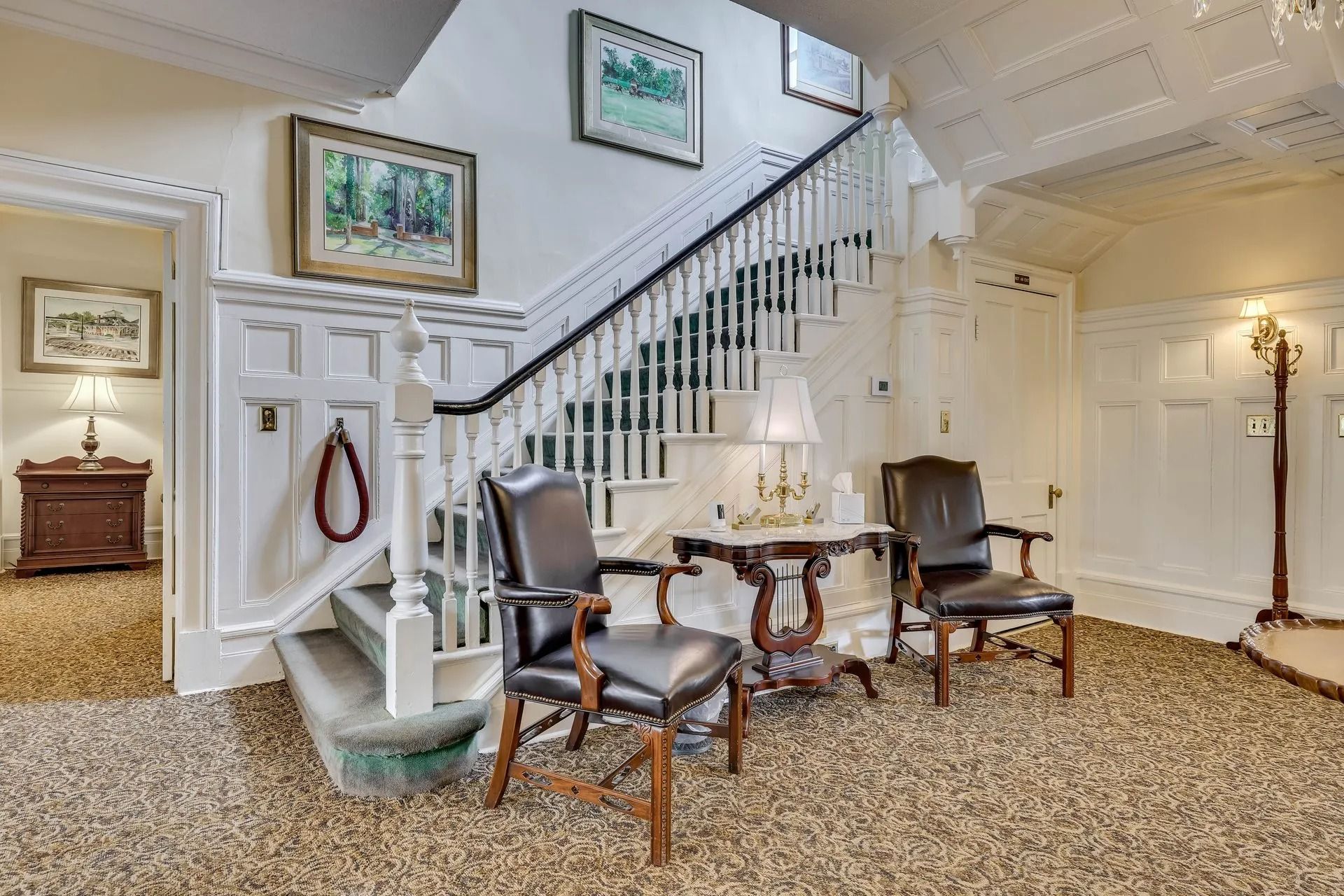 An entry hall with a staircase, two dark leather chairs, a small table, and framed art on white paneled walls.