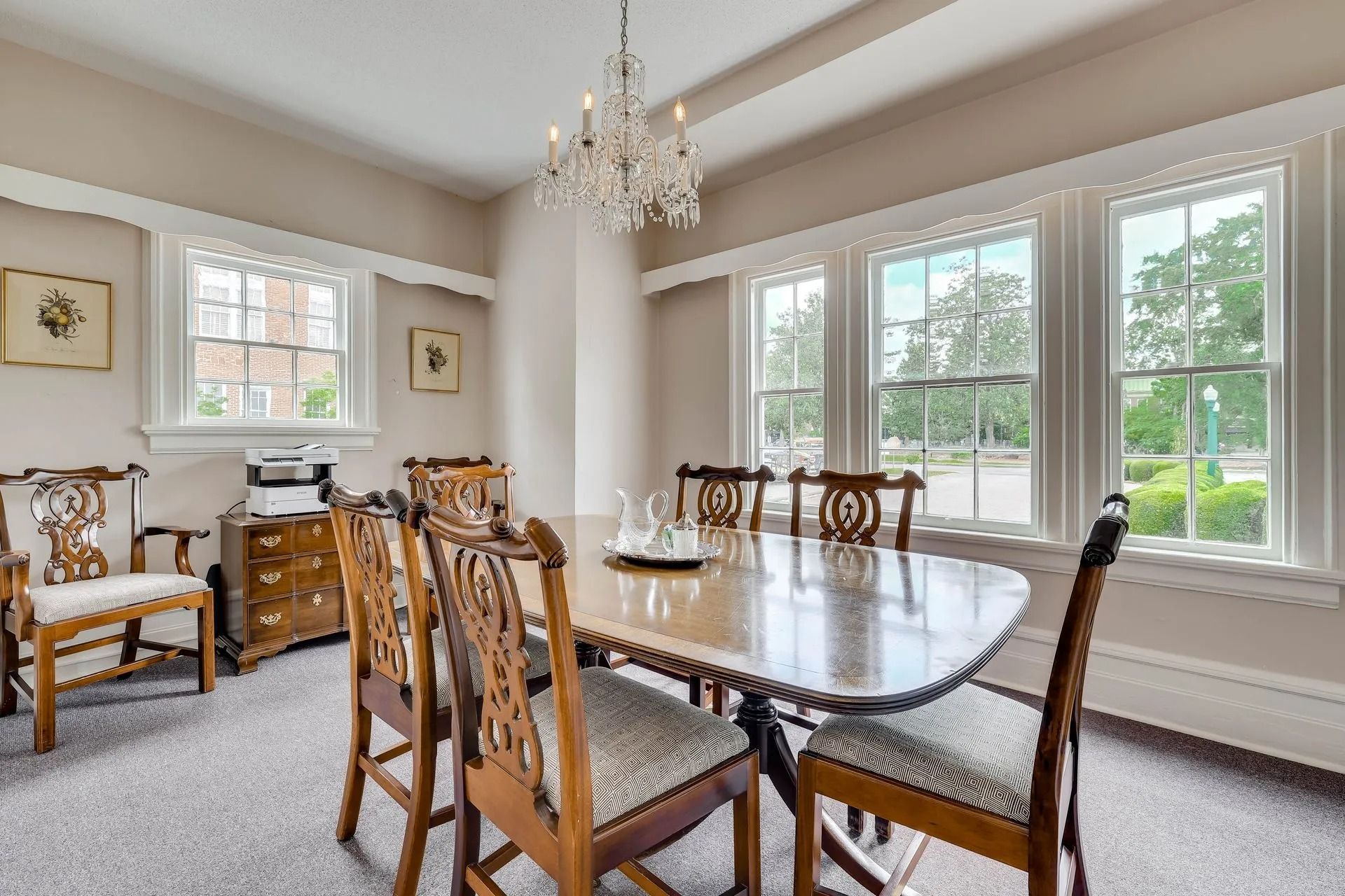 A dining room with a long wooden table, six chairs, a crystal chandelier, and large windows with light-colored walls.