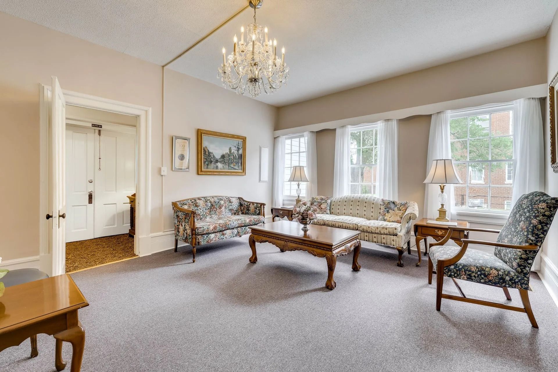 A formal sitting room with a chandelier, two floral sofas, a central coffee table, and an armchair on a carpeted floor.
