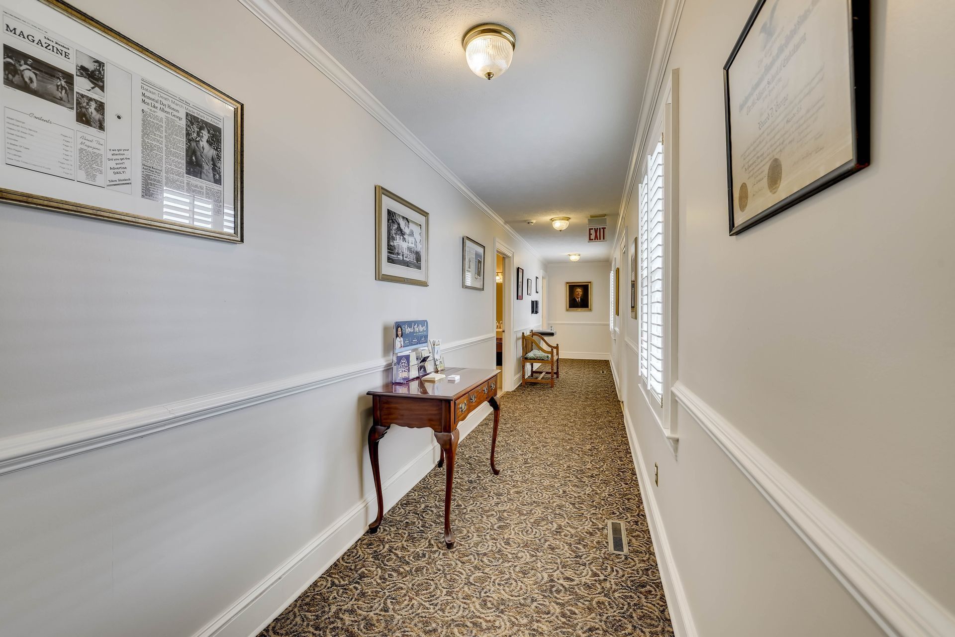 A brightly lit, beige-walled hallway with patterned carpet, a wooden side table, and framed pictures along the walls.