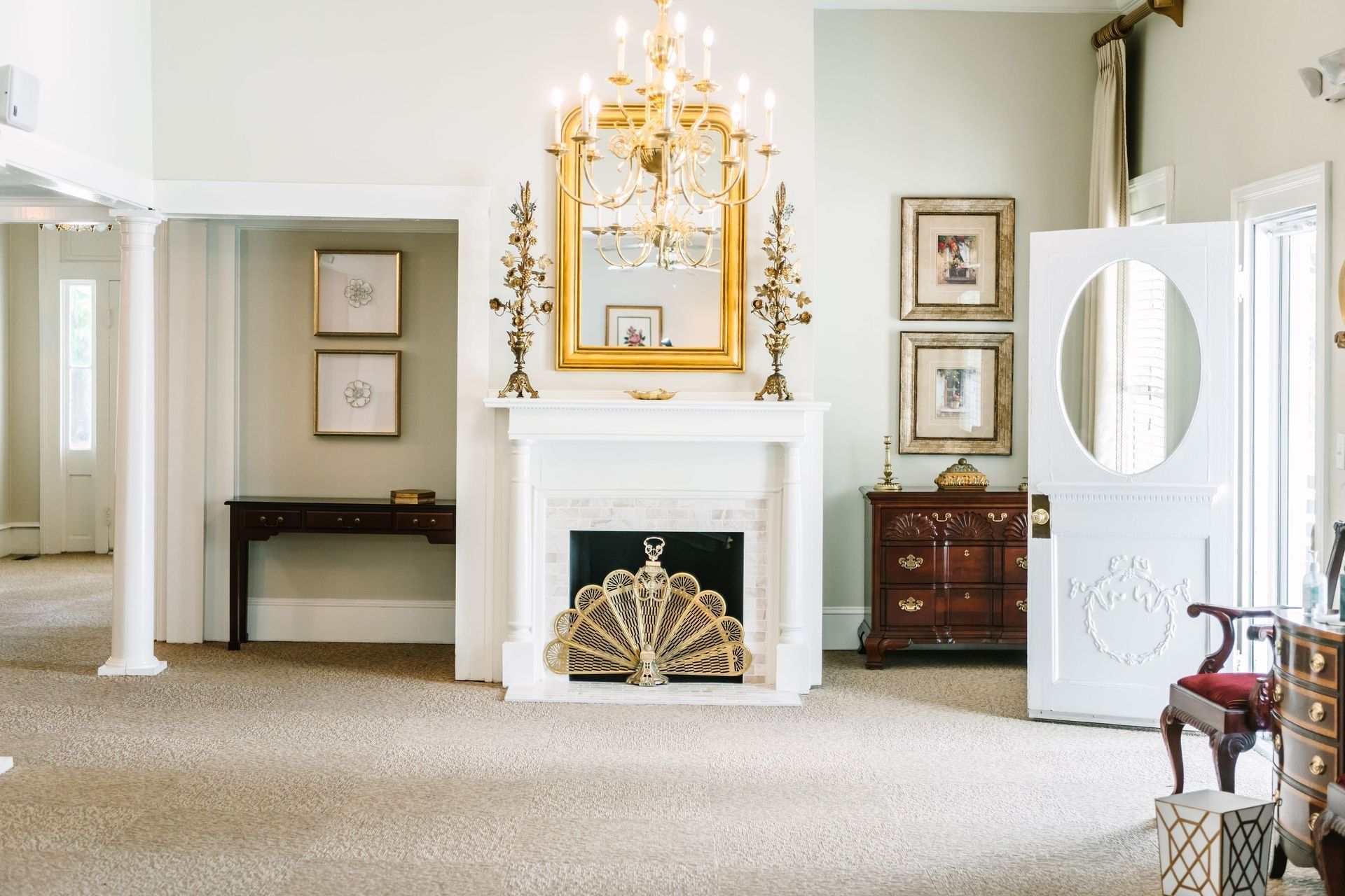 A formal living room with a fireplace, chandelier, ornate gold mirror, wood furniture, and a white door with an oval window.