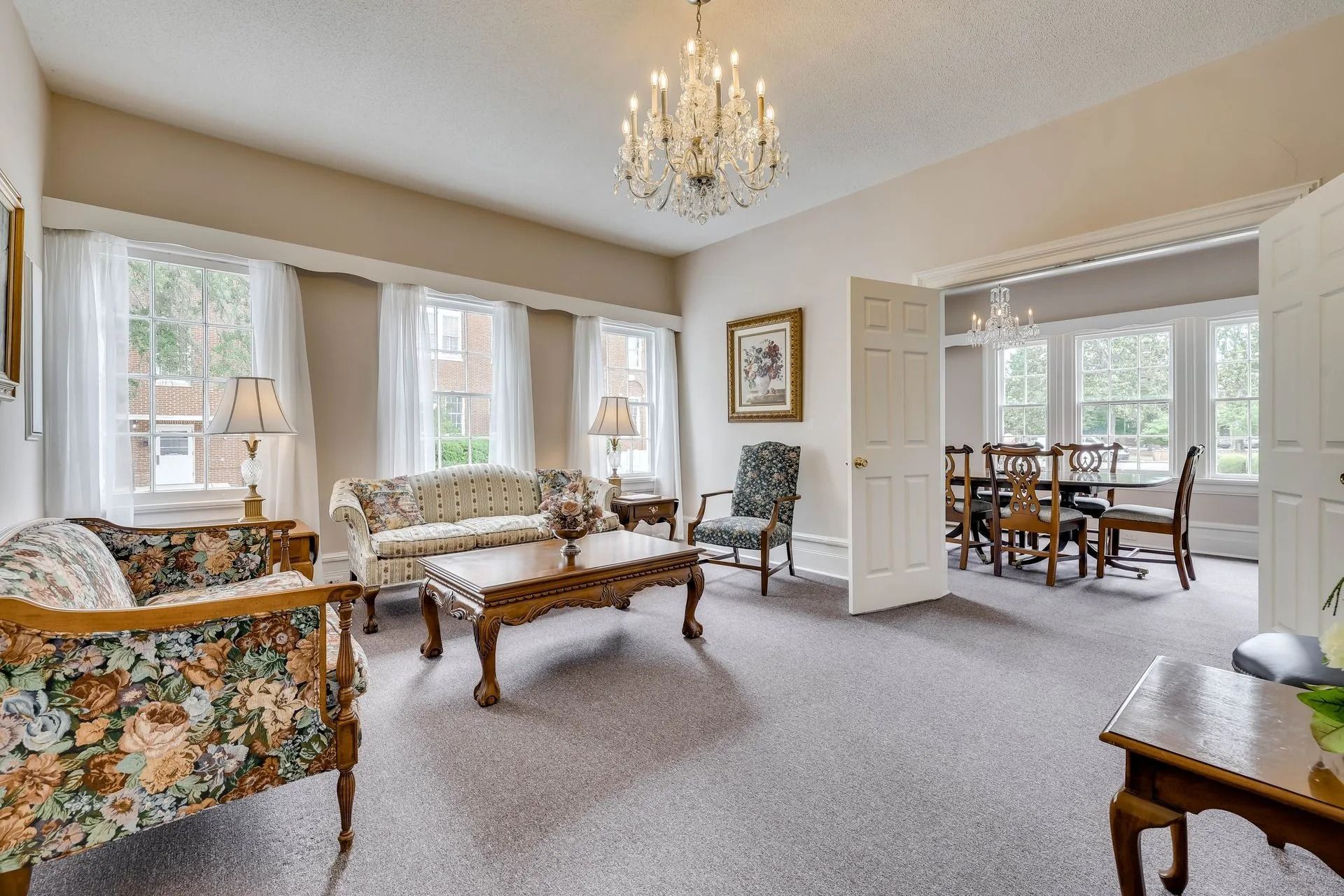 A furnished living room with a chandelier, floral sofa, coffee table, and an open doorway to a dining room.