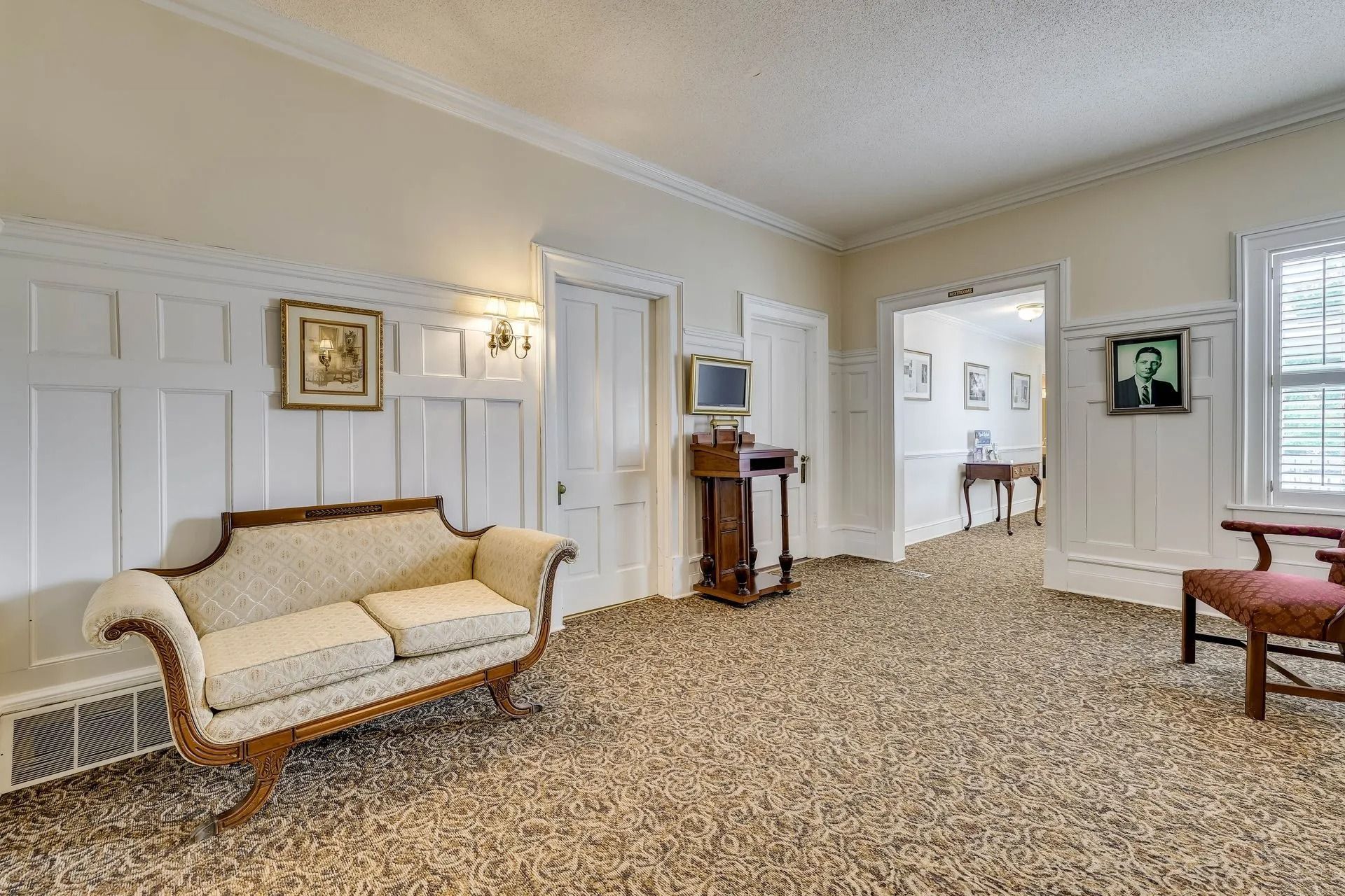 A formal room with white paneled walls, a cream settee, a wooden stand, and a red chair on patterned wall-to-wall carpet.