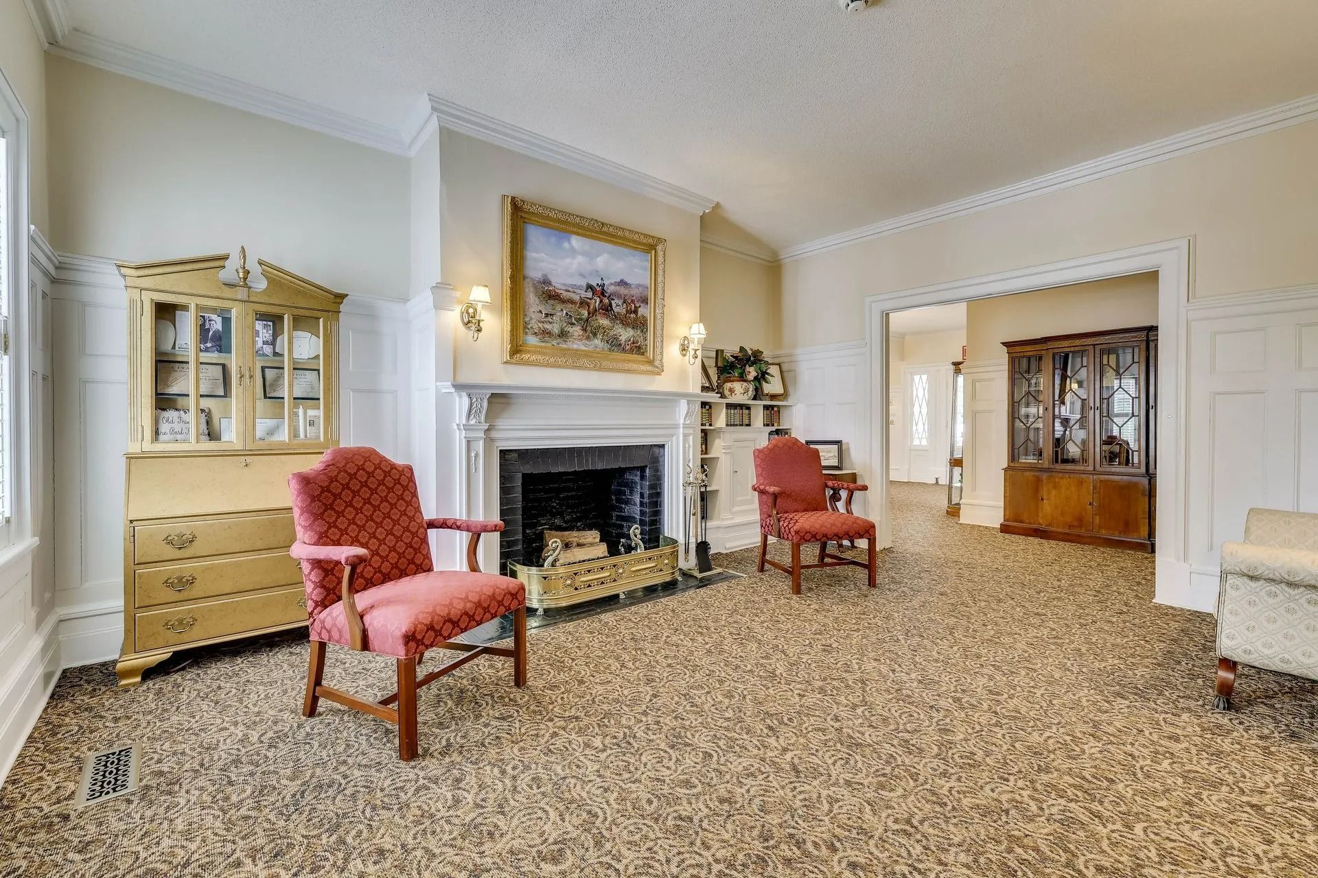 A formal living room with two red chairs facing a fireplace, a gold cabinet, and a wooden hutch in the background.