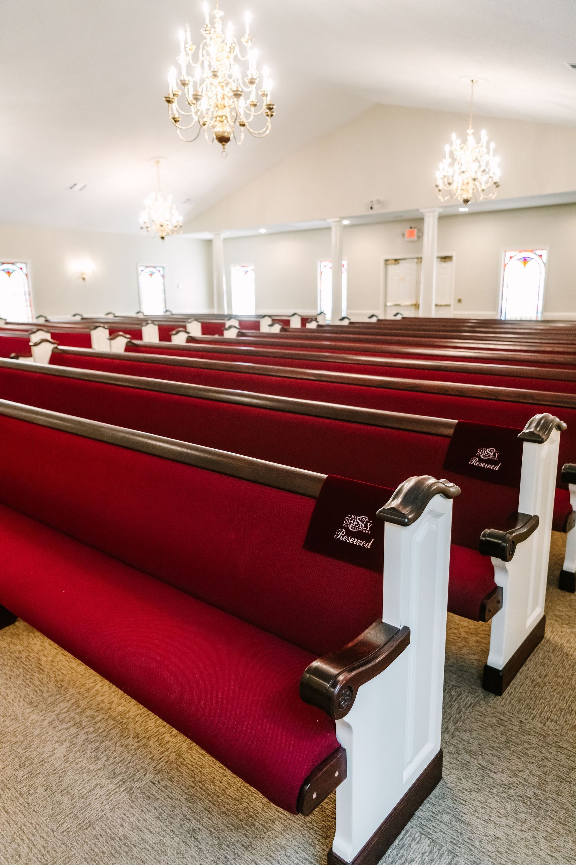 Rows of pews with red cushioned seats and white wooden ends inside a bright, empty church hall with crystal chandeliers.