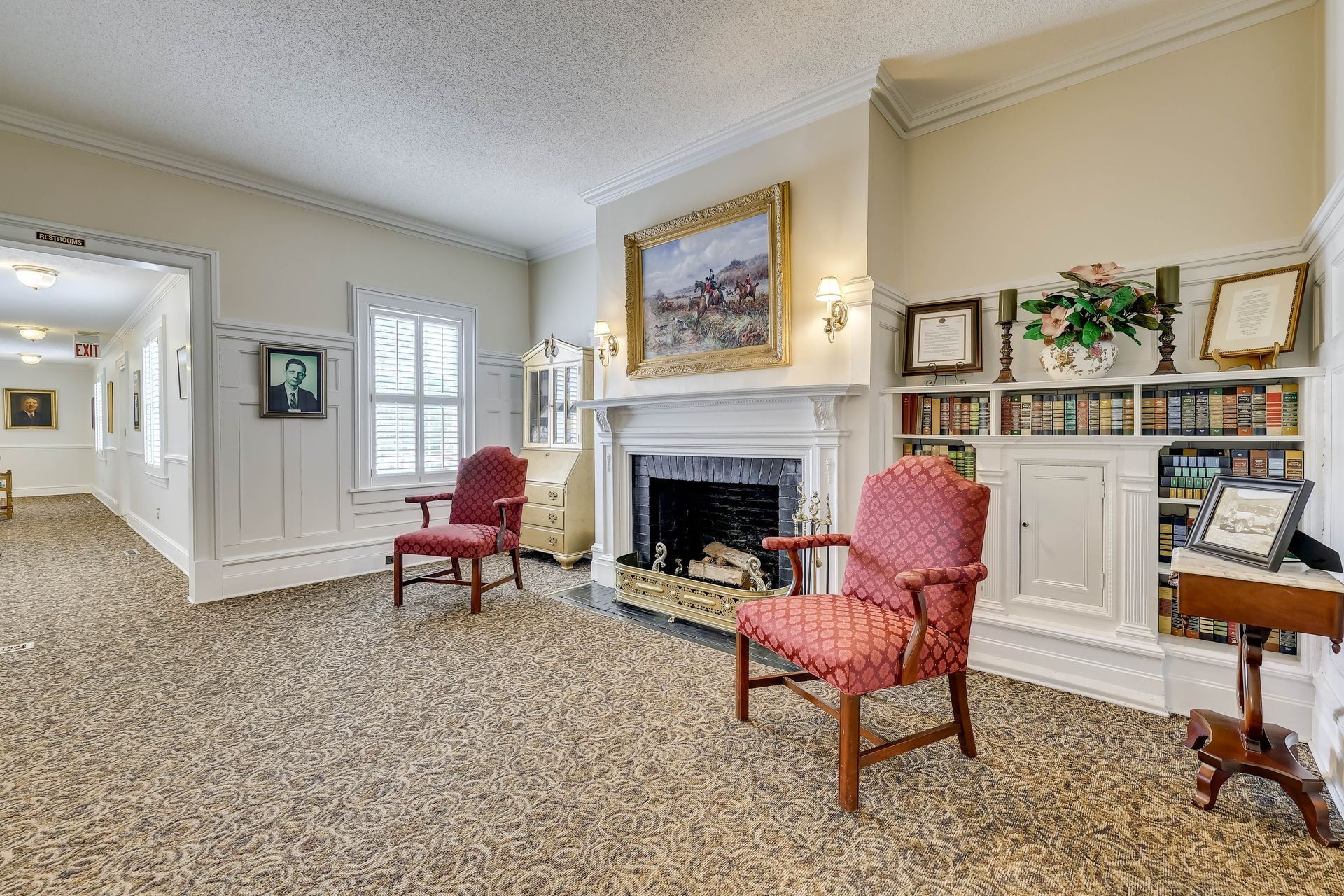 A sunlit room features a fireplace with a framed painting, two red armchairs, a bookshelf, and an open hallway.