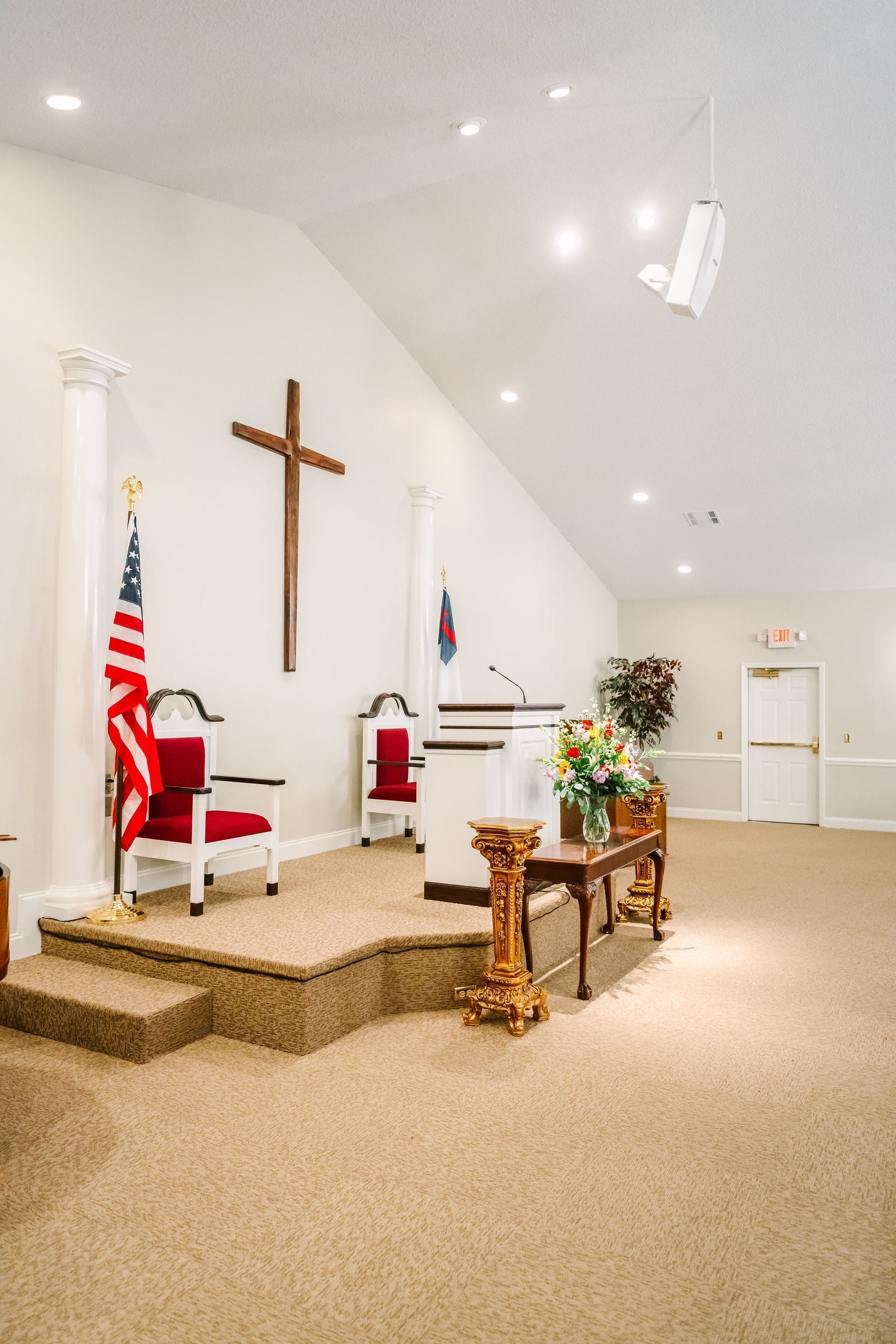 Interior of a church sanctuary featuring an altar, a wooden cross on the wall, an American flag, and floral arrangements.