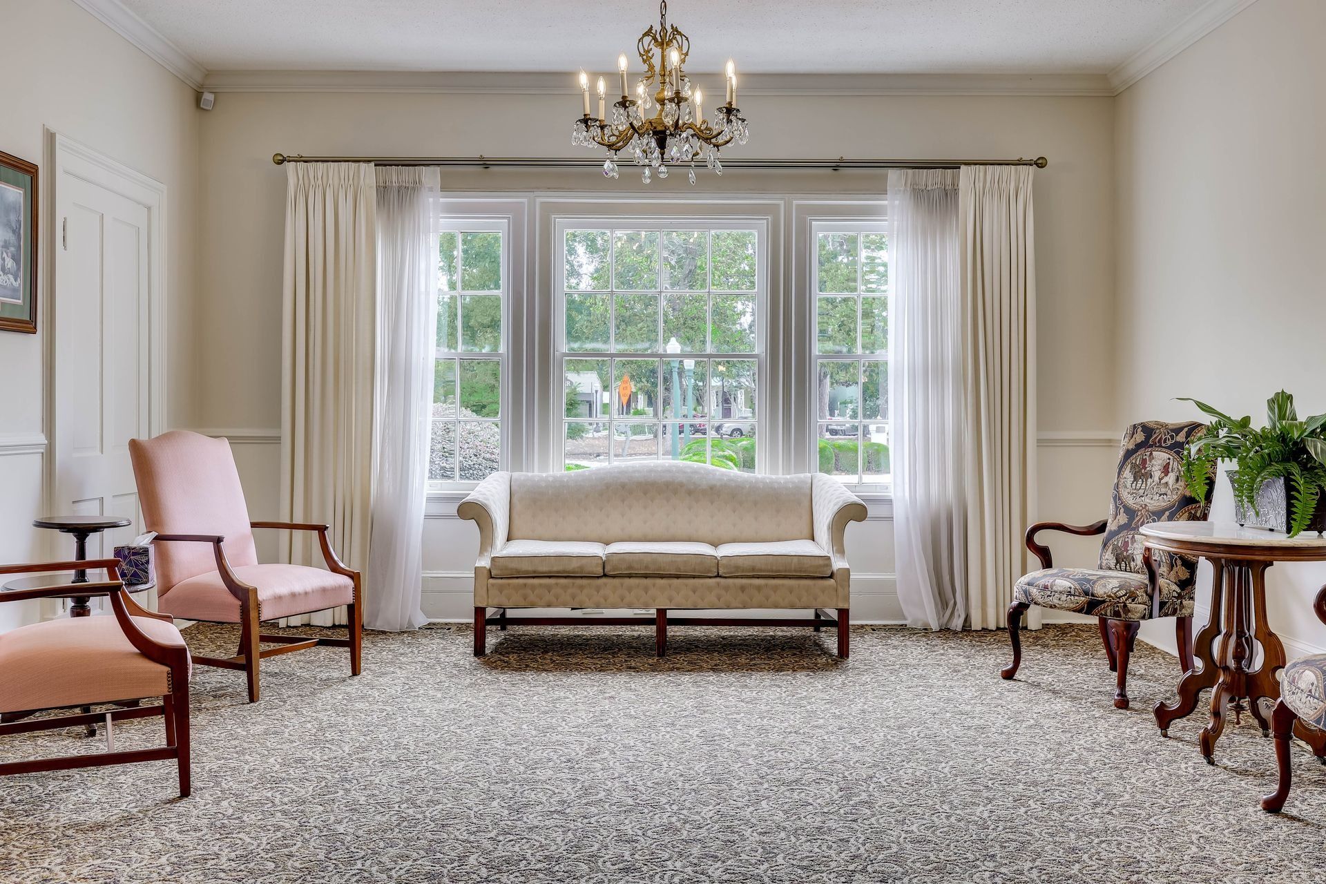 A bright, neutral-toned living room featuring a centered sofa, two armchairs, a side table with a plant, and a chandelier.