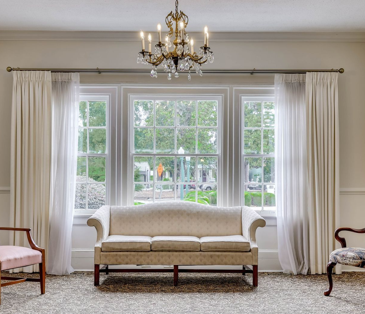 A classic living room with a neutral sofa beneath a triple-pane window, draped in floor-length curtains and a chandelier.